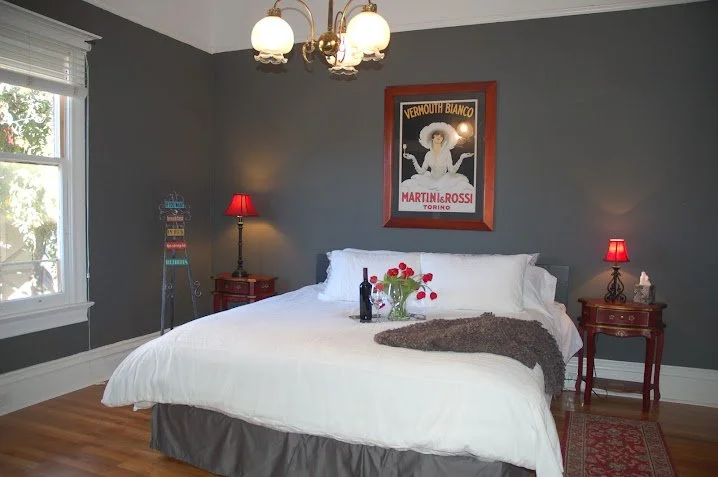 Bedroom with gray walls, a large bed with white bedding and pillows, a dark brown blanket, and a vase with red flowers on top. Two red bedside lamps and a vintage-style poster on the wall above the bed. Two windows with white trim, hardwood floors, and a ceiling light fixture with multiple bulbs.