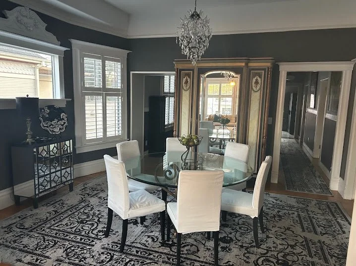 Dining room with six white upholstered chairs around a glass-top table, a decorative chandelier hanging above, dark patterned rug on the floor, antique-style armoire, window with white shutters, and a side table with a lamp and decorative pieces.