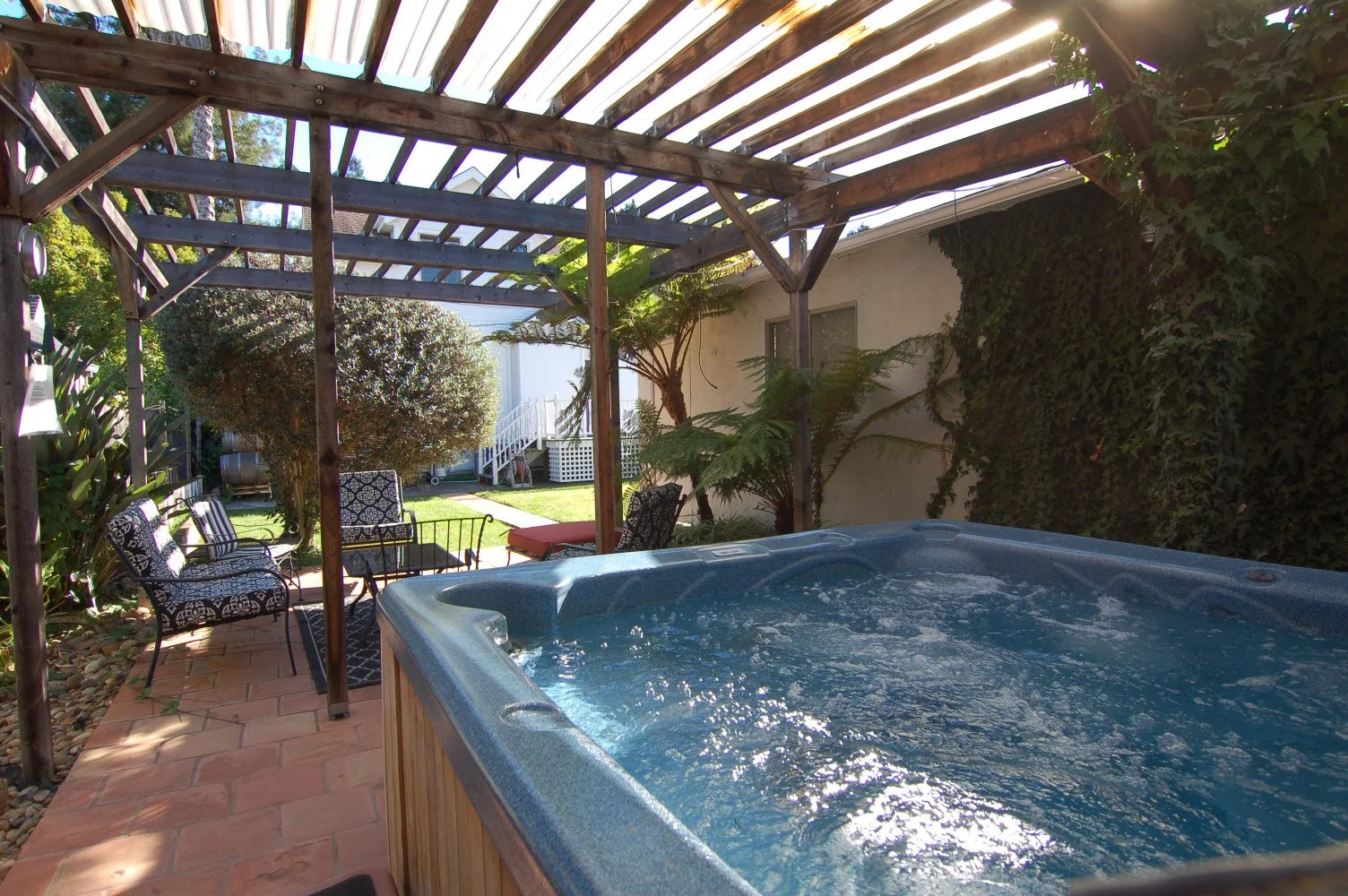 Outdoor backyard with hot tub, patio furniture, and a pergola, surrounded by greenery and plants.