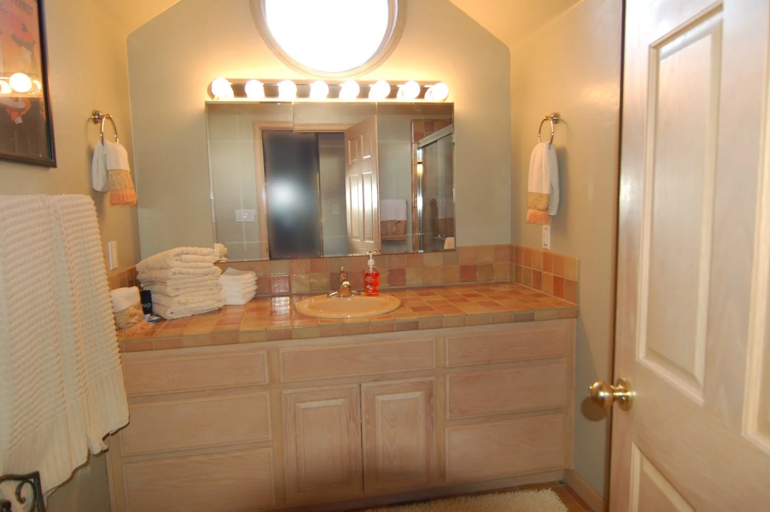 Bathroom vanity with a large mirror, stack of towels, sink with soap dispenser, surrounded by tiled countertop and cabinets, with towels hanging on either side and a door to the right.