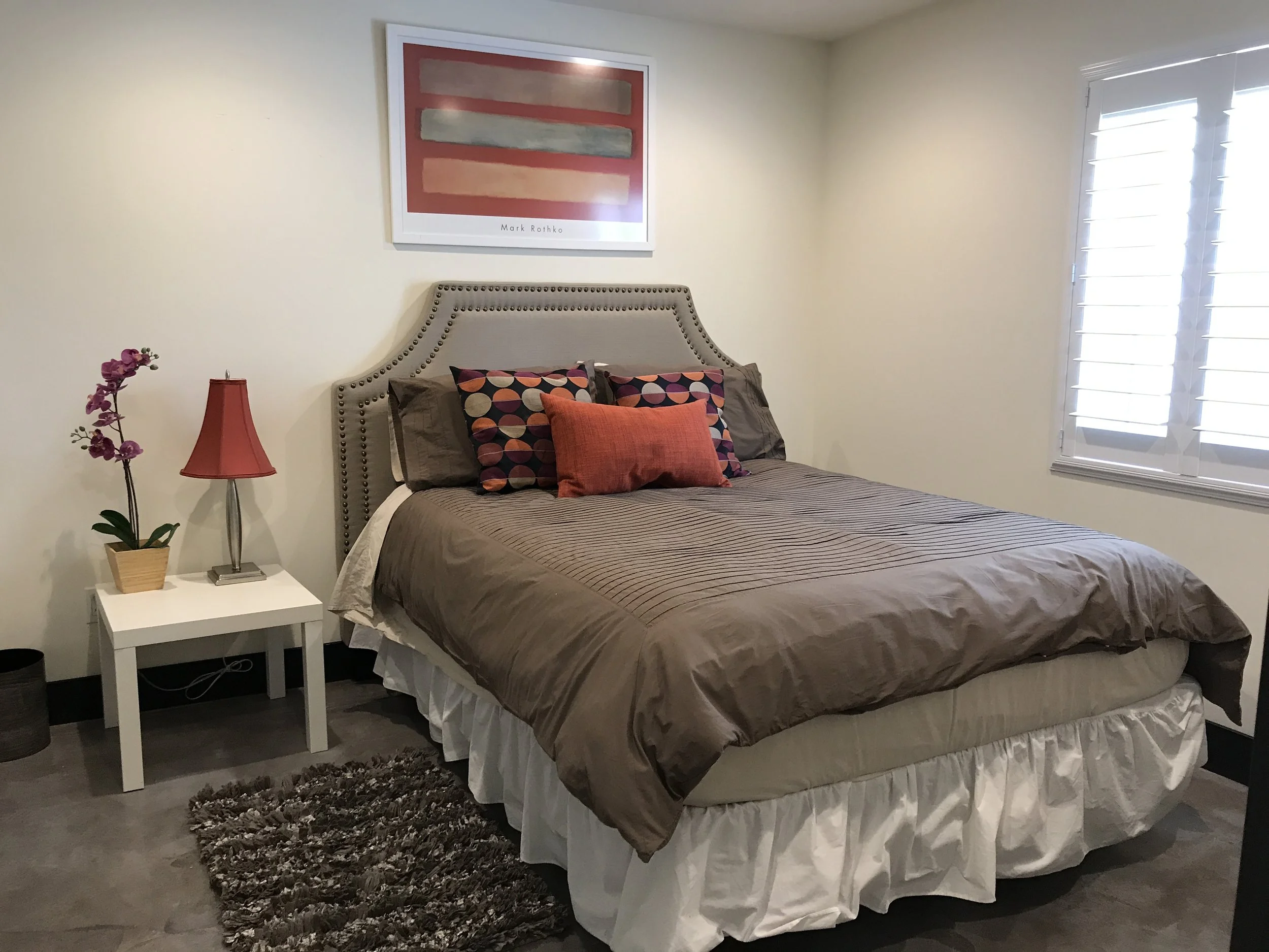A bedroom with a bed that has a gray upholstered headboard, beige bedding with striped pattern, and decorative pillows in circles and coral colors. There is a white nightstand with a red lamp and purple orchid plant. A framed abstract art piece hangs above the bed. The room has cream walls, a window with white shutters, and a brown shaggy rug next to the bed.