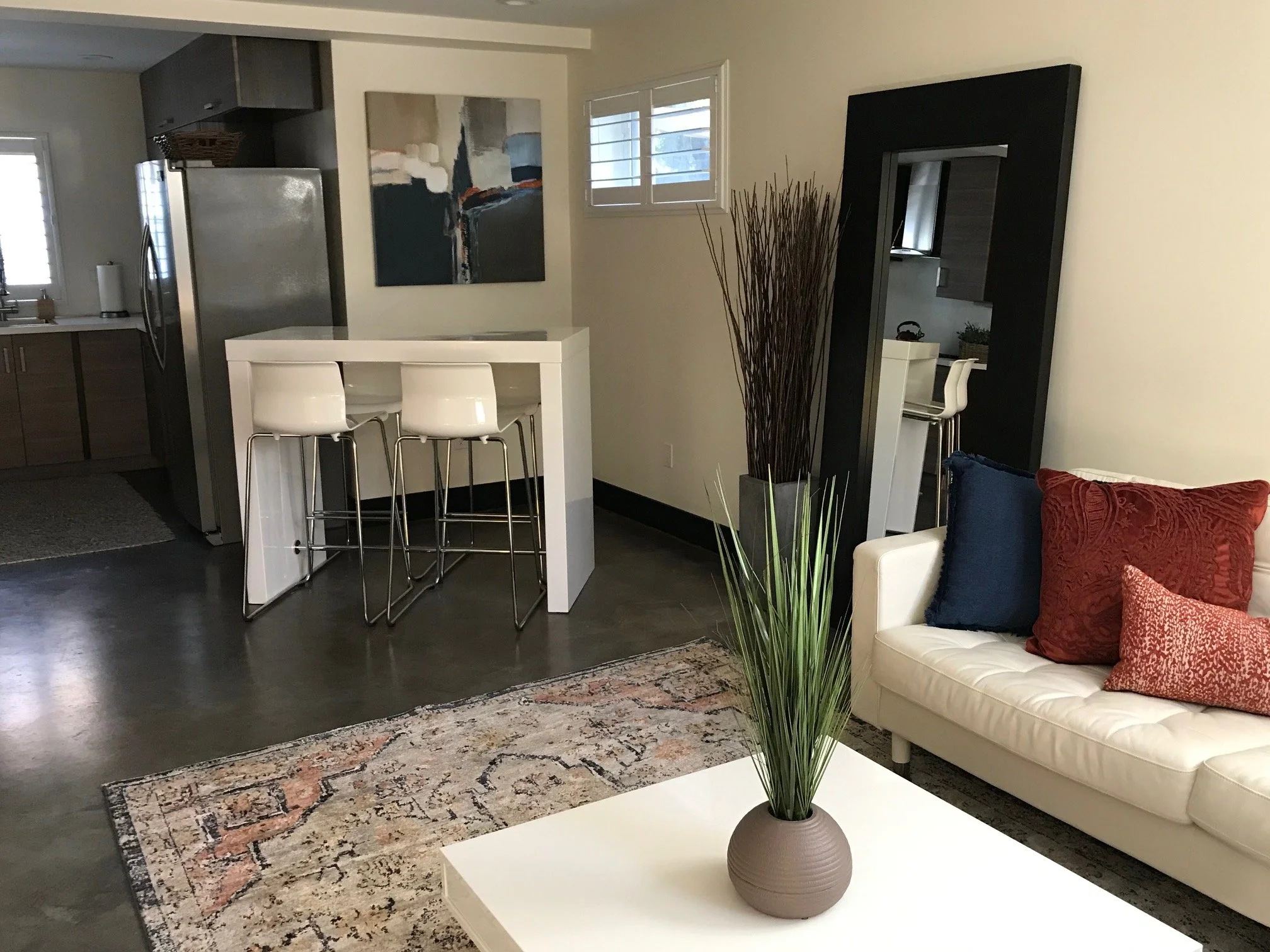 Living room with a white sofa, decorative pillows, a coffee table with a potted plant, a large black-framed mirror, and kitchen area in the background with bar stools and abstract artwork.