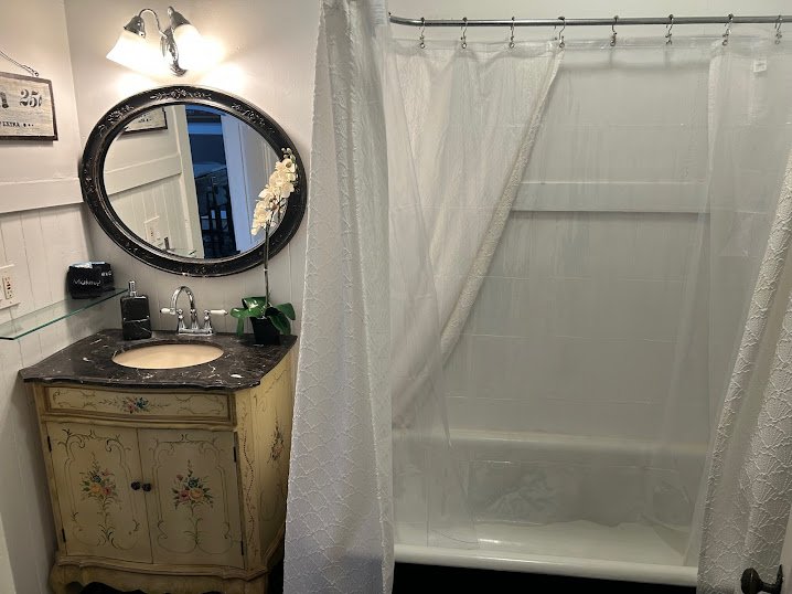 Small bathroom with a vintage wooden vanity, black countertop, and oval mirror. A shower with white curtains is visible on the right. The wall has white paneling, and there is a framed picture or sign above the vanity.