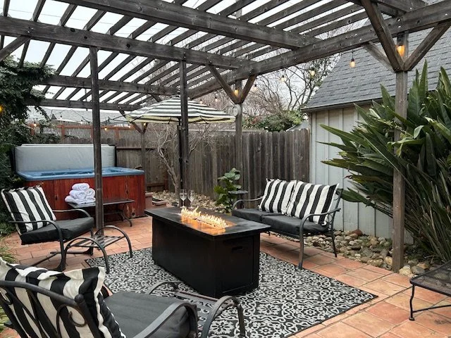 Outdoor patio with seating area, fire pit table, hot tub, umbrella, string lights, rock landscaping, and a porch railing with trees in the background.