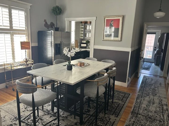 Modern dining room with a white marble table surrounded by upholstered chairs, a rug beneath the table, a framed artwork on the wall, a stainless steel refrigerator, and a sideboard with decorative items. Natural light from windows on the left.