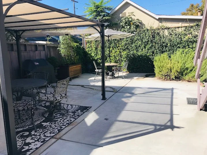 A backyard patio with concrete flooring, covered seating area with black metal chairs and a table, a BBQ grill, and an outdoor table with chairs under a large umbrella. There are lush green bushes and a partially visible house in the background.