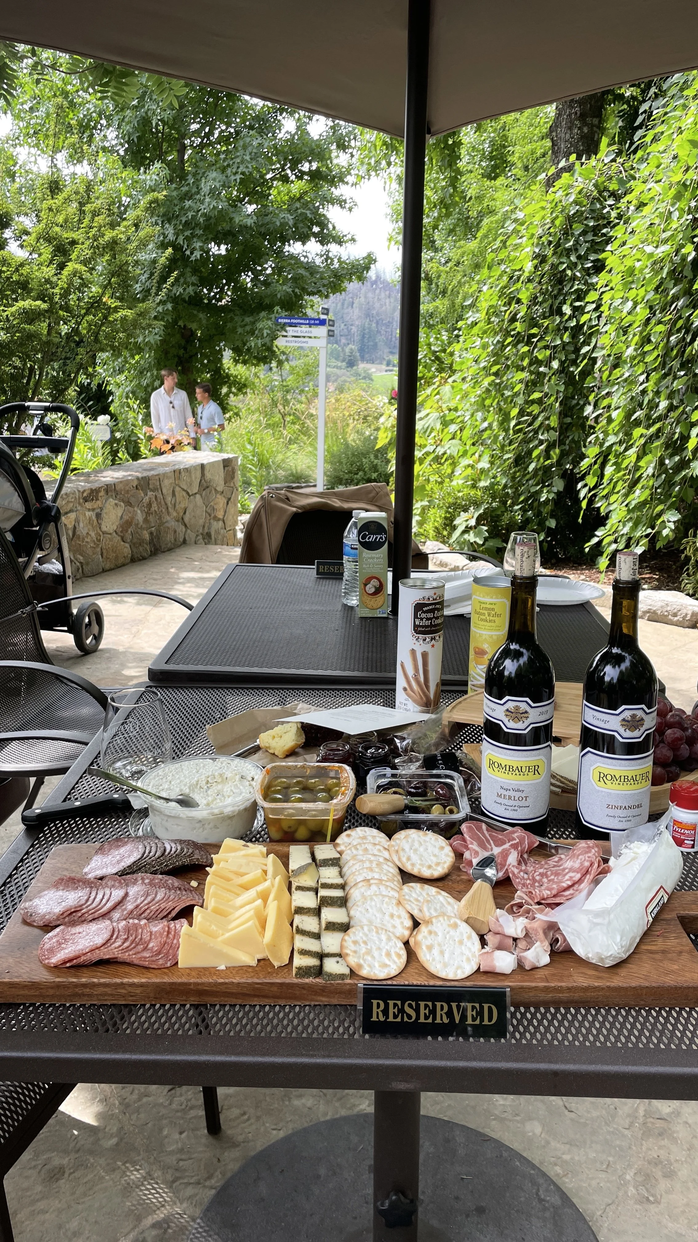 A reserved table at an outdoor gathering with a cheese and charcuterie platter, two bottles of red wine, grapes, and condiments, set under a large umbrella on a patio surrounded by greenery, with two people talking in the background.