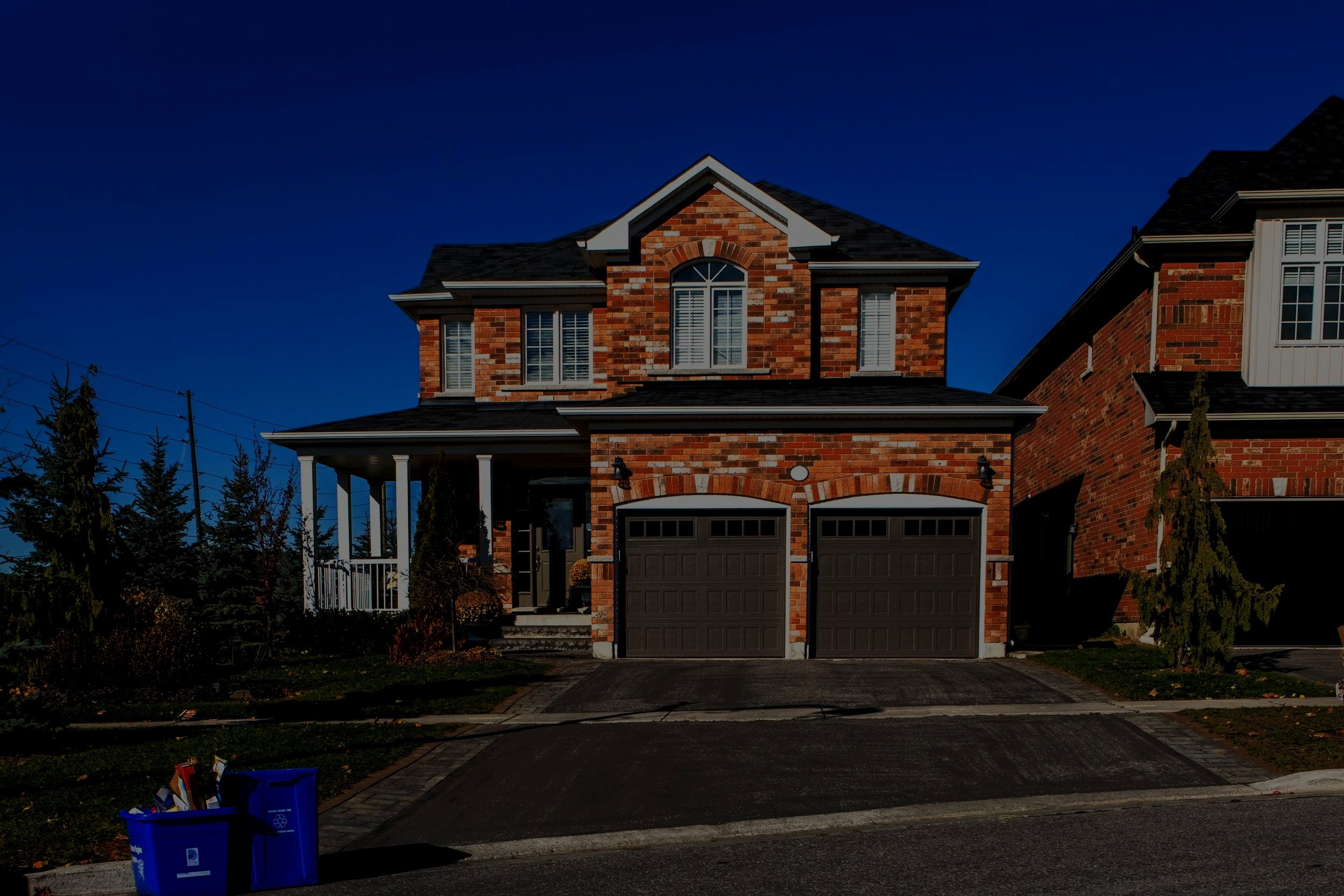 A two-story brick house with a dark shingle roof and two black garage doors, set against a clear blue sky. There is a porch with white columns on the left side, some trees in the front yard, and trash bins at the driveway edge.