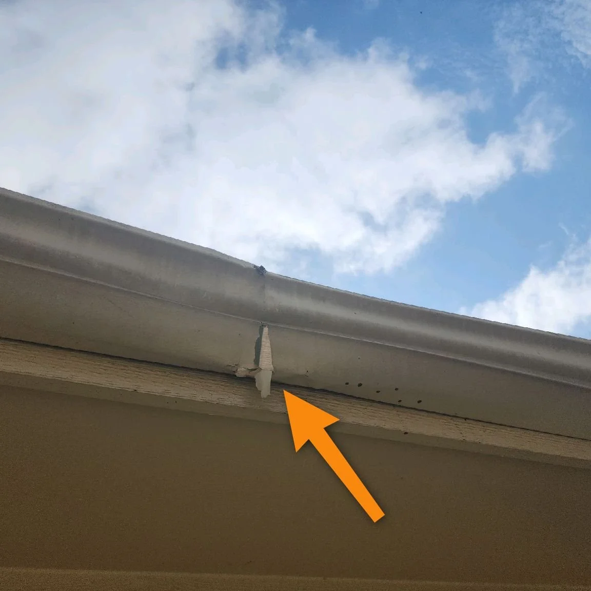 Close-up view of a damaged section of an exterior house gutter and soffit, with an orange arrow pointing to the damaged area, against a background of a blue sky with clouds.