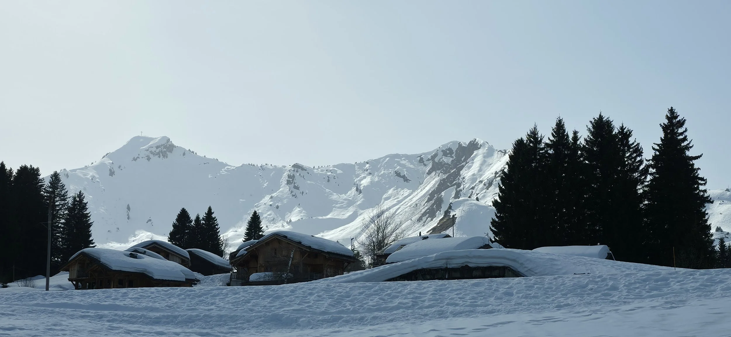 Paysage de montagne enneigée avec des chalets en bois, des sapins et des montagnes en arrière-plan sous un ciel clair.