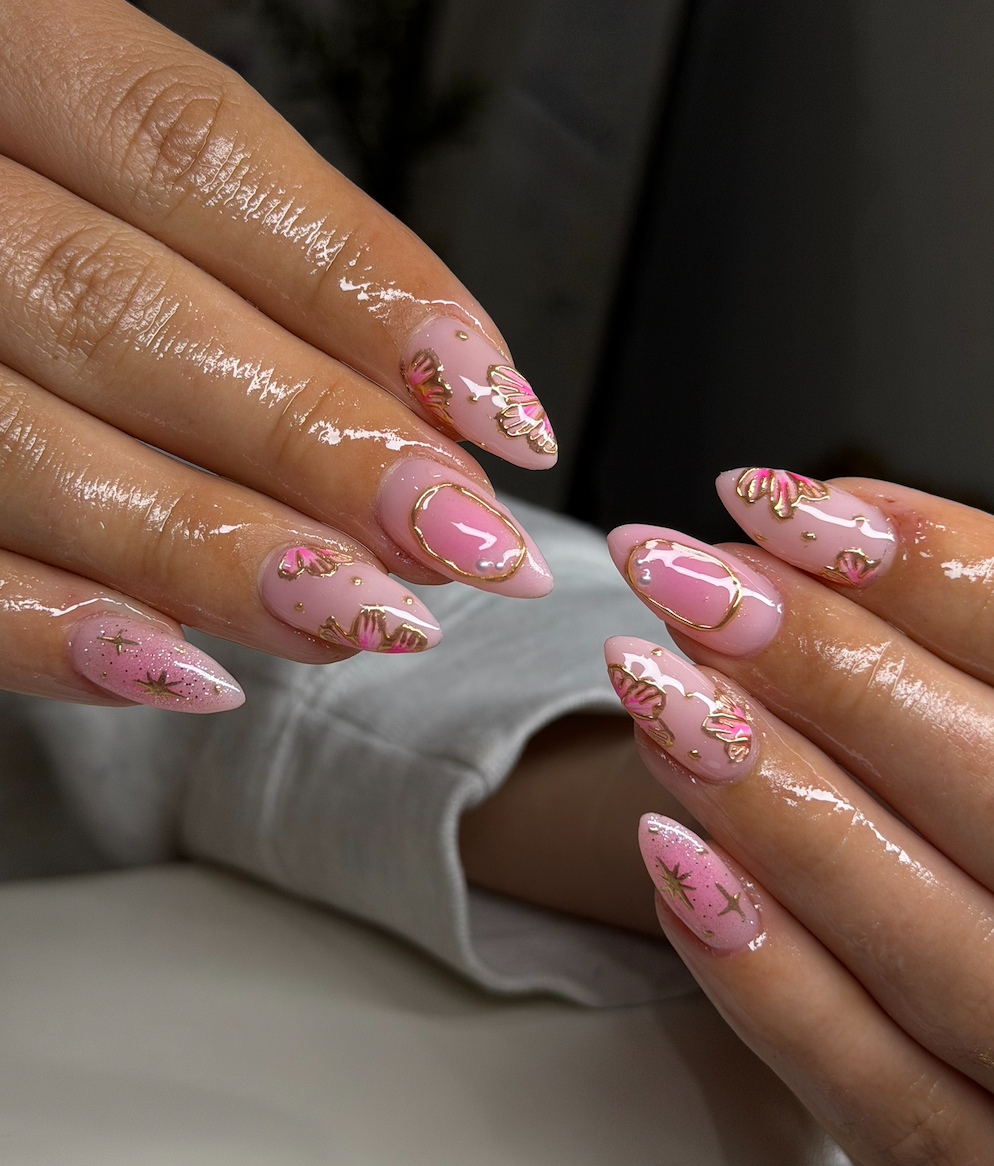 Close-up of hands with manicured nails featuring pink and gold nail art, including floral designs, glitter, and stars.