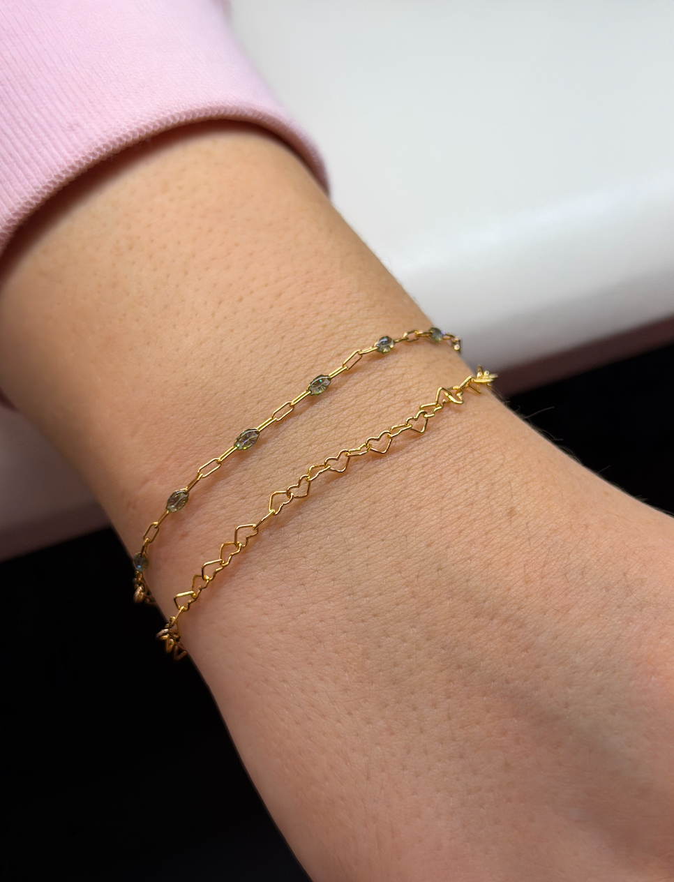 Close-up of a wrist with two gold bracelets, one with small gemstone accents and the other with a heart and zigzag pattern, against a pink shirt cuff and light background.