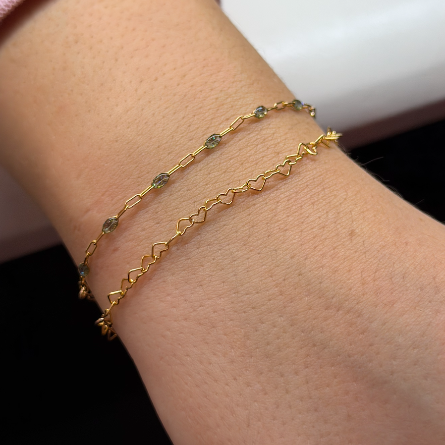 Two gold bracelets, one with small gemstones and the other with a heart and zigzag design, worn on a person's wrist.