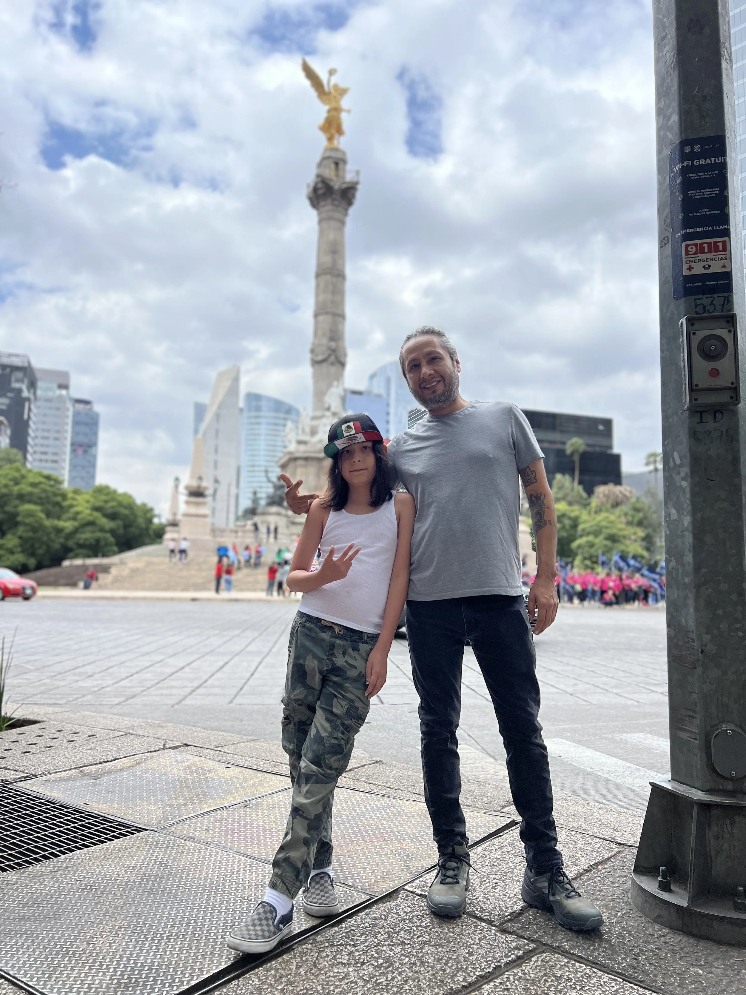 A man and girl posing in front of a monument with a golden statue on top, in an urban area with modern buildings, trees, and people in the background.