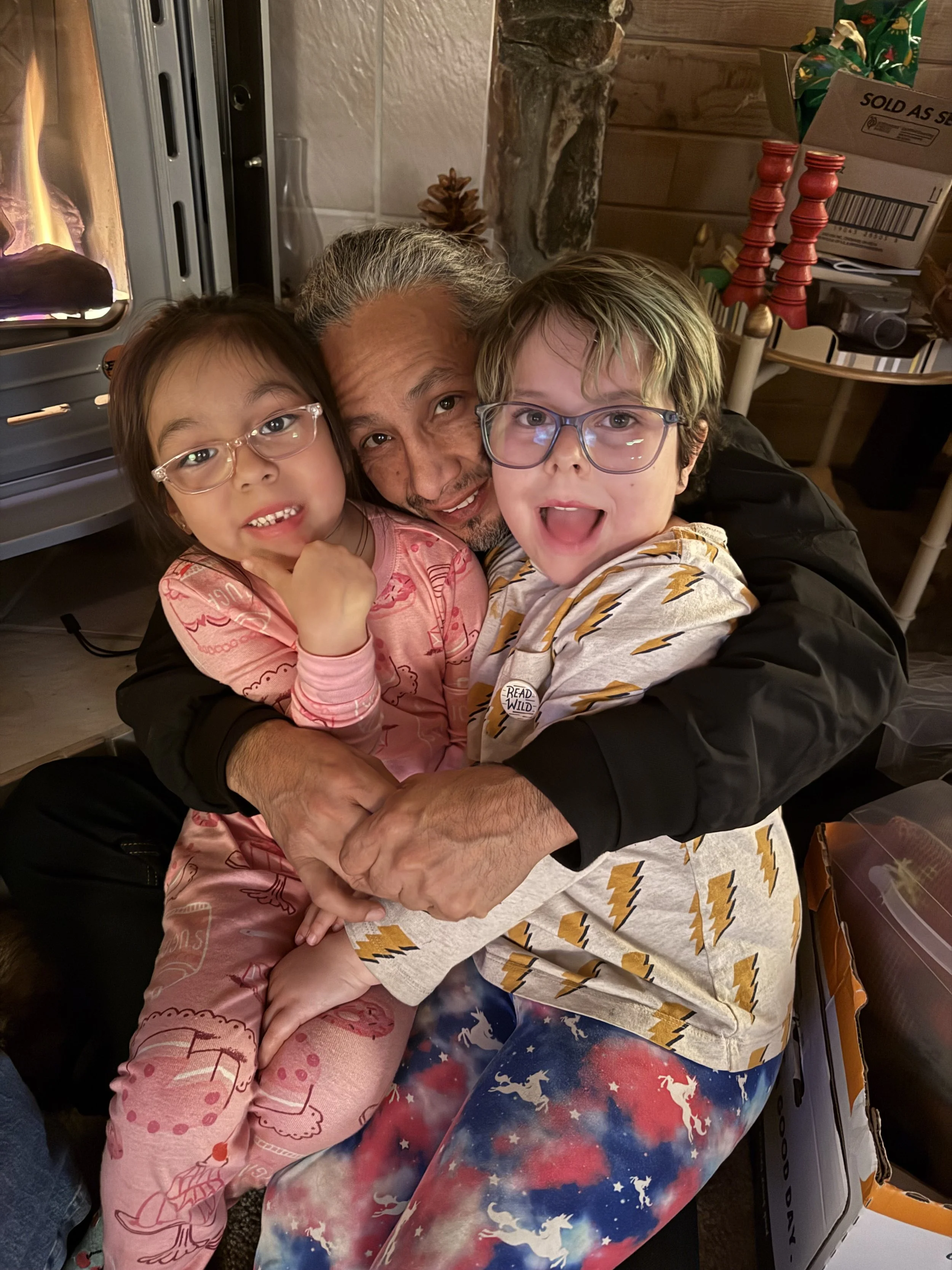 A man hugging and playing with two young children, one girl and one boy, near a fireplace with a cozy indoor setting.