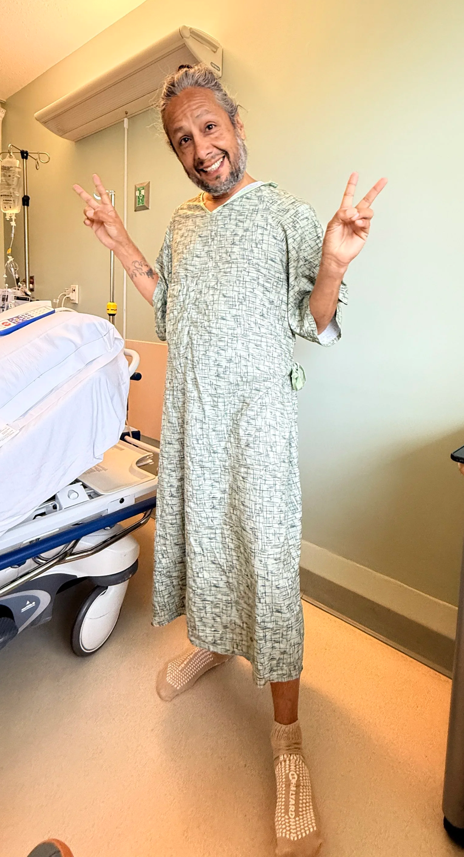 Happy man in hospital gown making peace signs with both hands in hospital room.