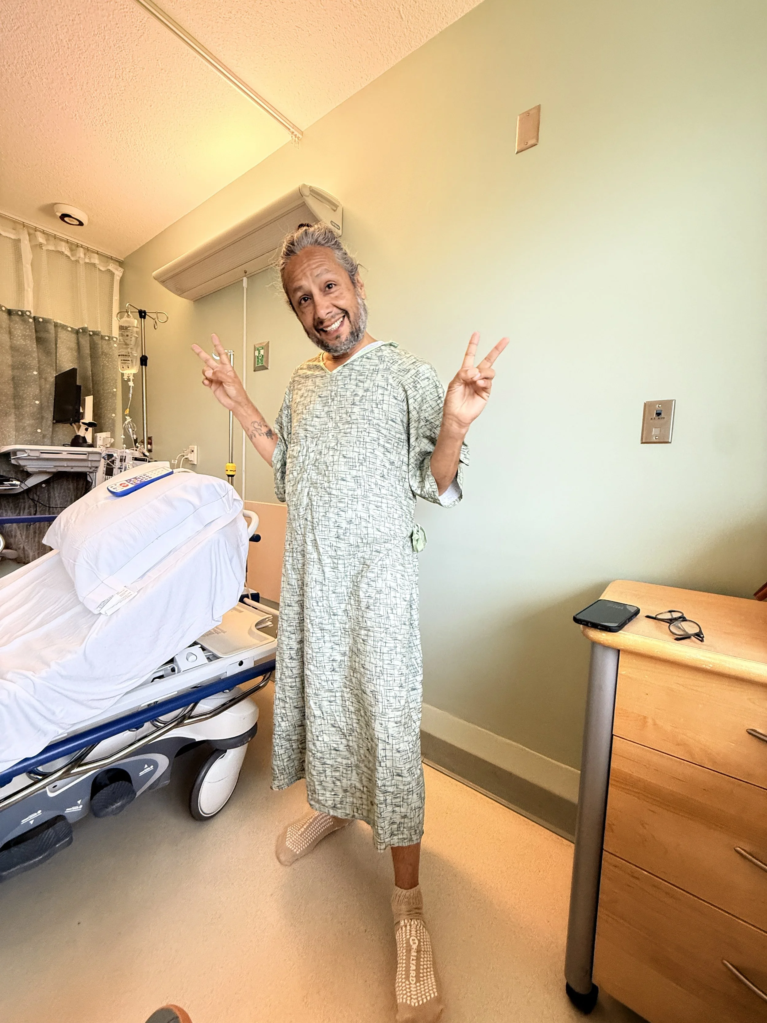 A man in a hospital gown making peace signs with both hands in a hospital room, standing near a hospital bed.