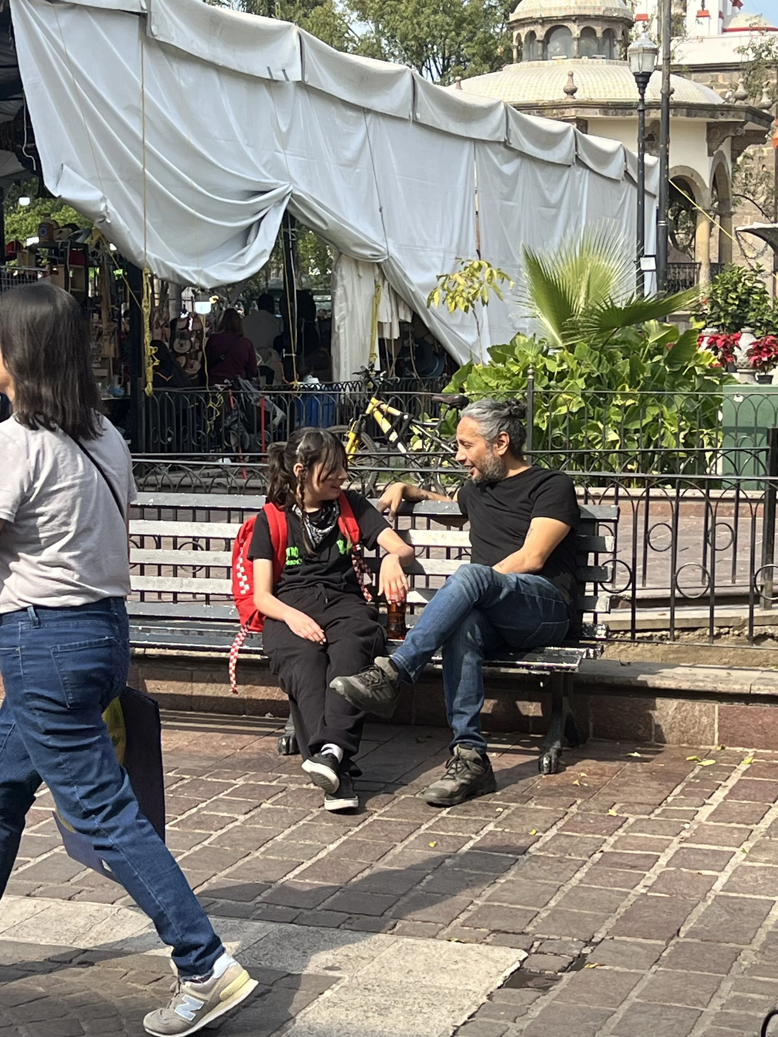 A man and a girl sit on a park bench, engaged in a conversation, with a woman walking past in the foreground. The girl has a red backpack and black clothing, while the man has gray hair, a black shirt, and jeans. There is a tent and greenery in the b