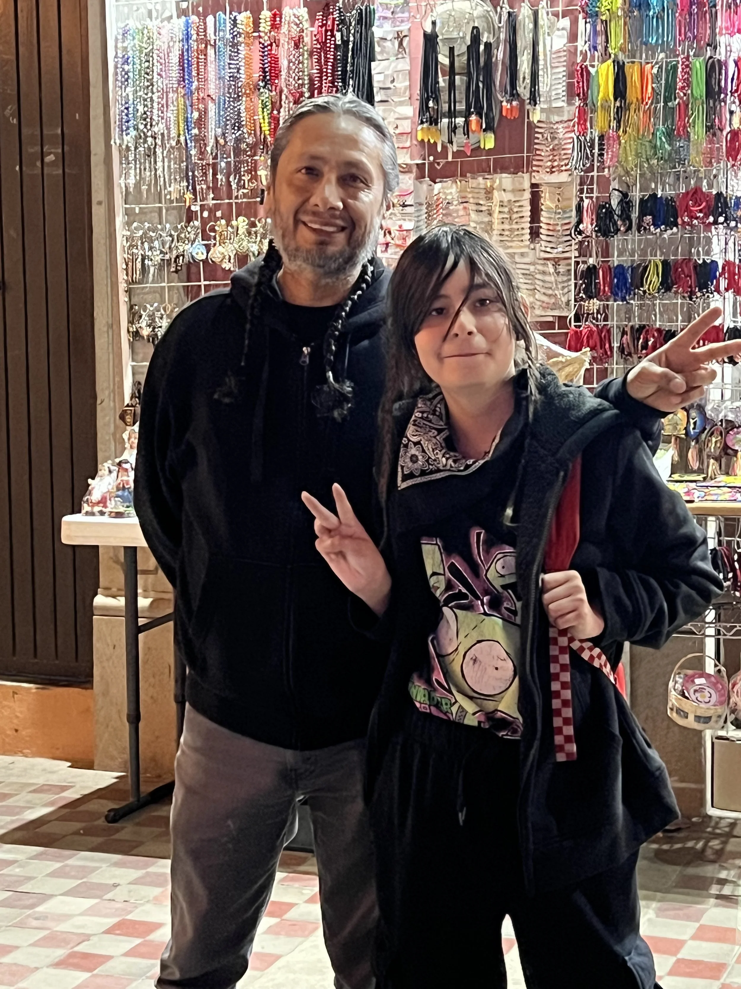 Two people standing inside a store with colorful accessories hanging on the wall behind them. The person on the left has gray hair, a beard, and is making a peace sign with their right hand. The person on the right has dark hair, is smiling, and maki