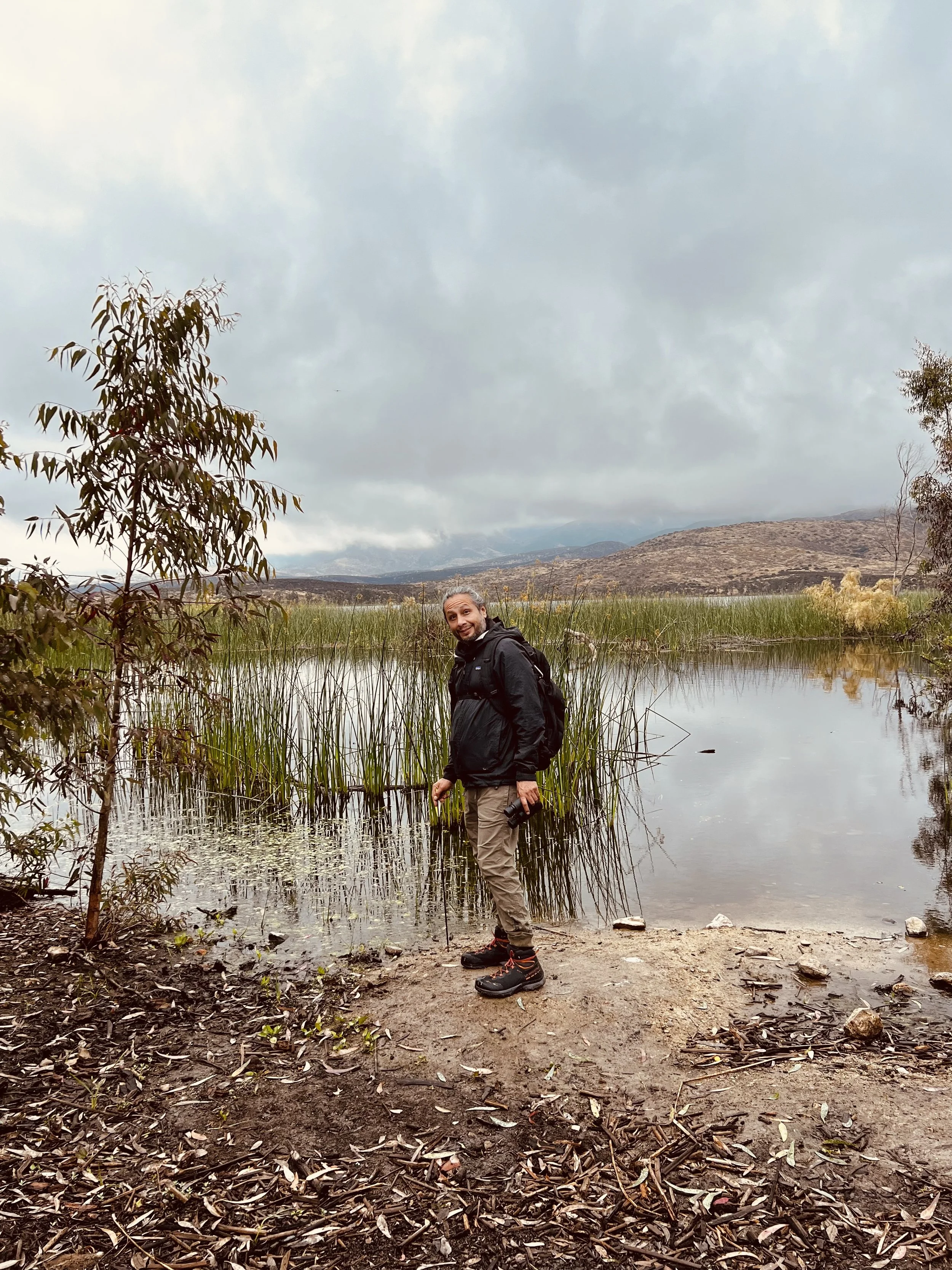 A man standing at the edge of a lake in a natural landscape with mountains and cloudy sky in the background.