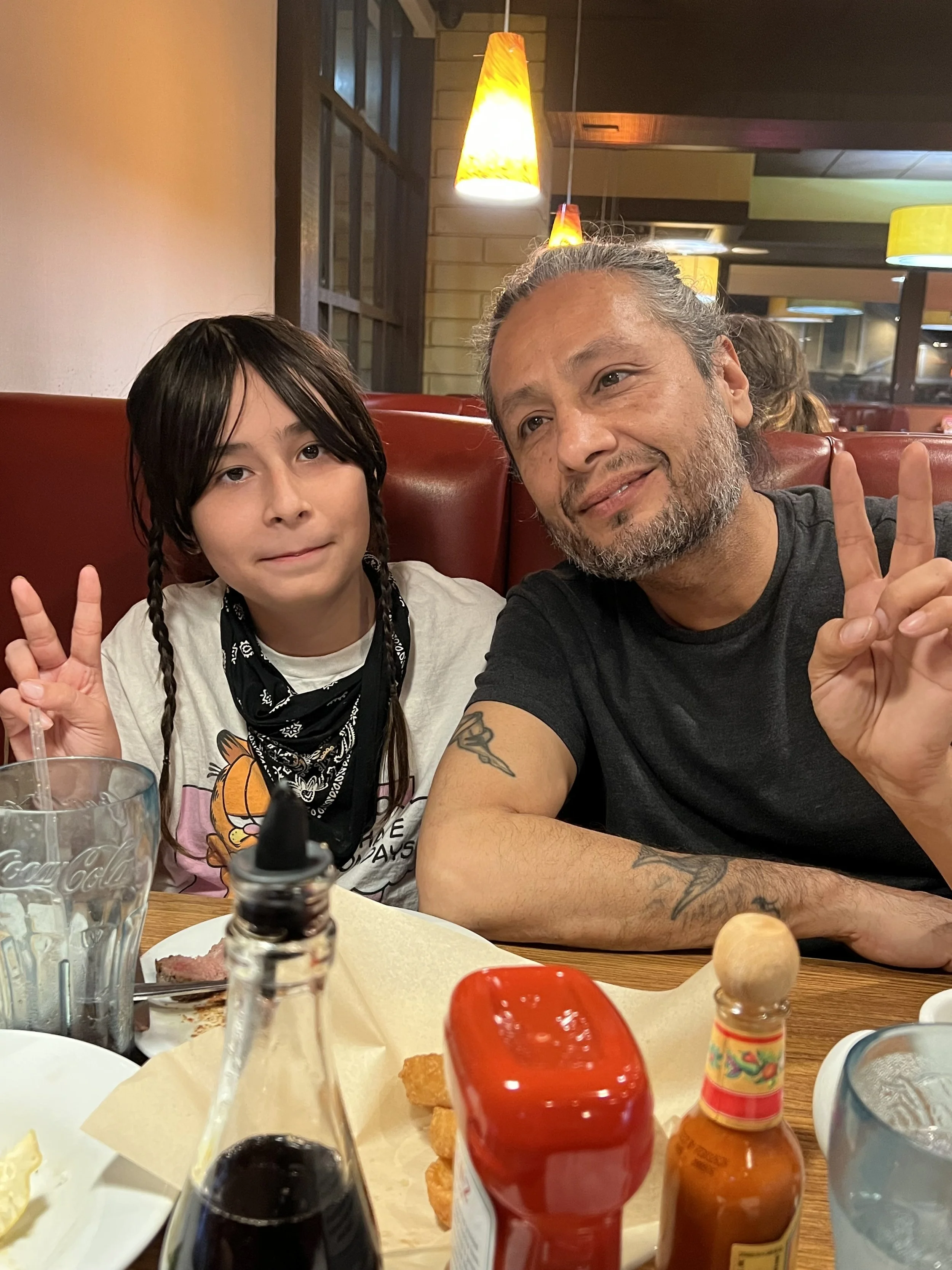 A man and a young girl sitting at a restaurant table, both giving peace signs. The table has condiments, a glass, and some food. The restaurant has warm lighting and a cozy ambiance.
