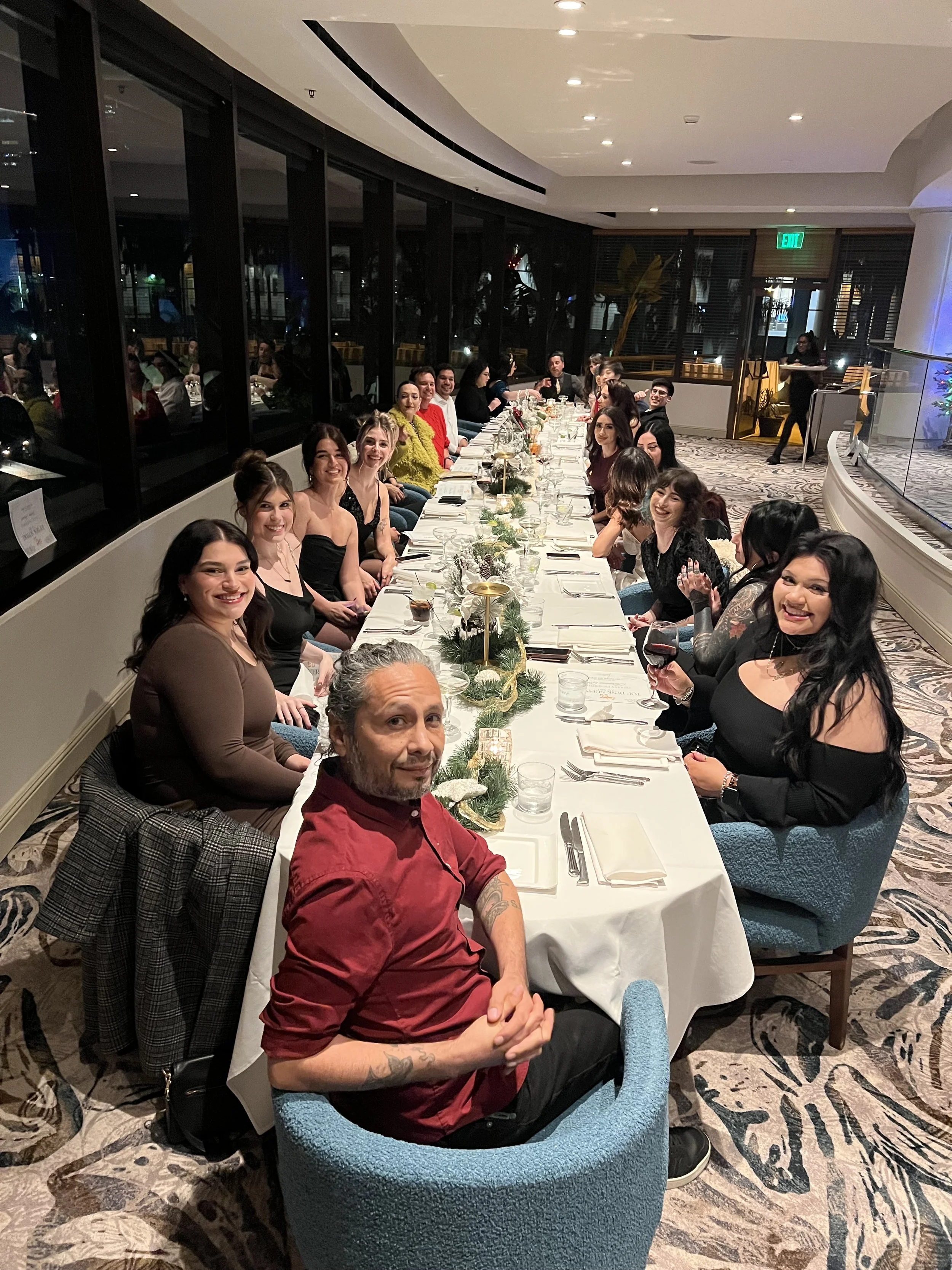 A group of people sitting at a long banquet table in a restaurant, smiling and enjoying a meal together, decorated with holiday-themed centerpieces.