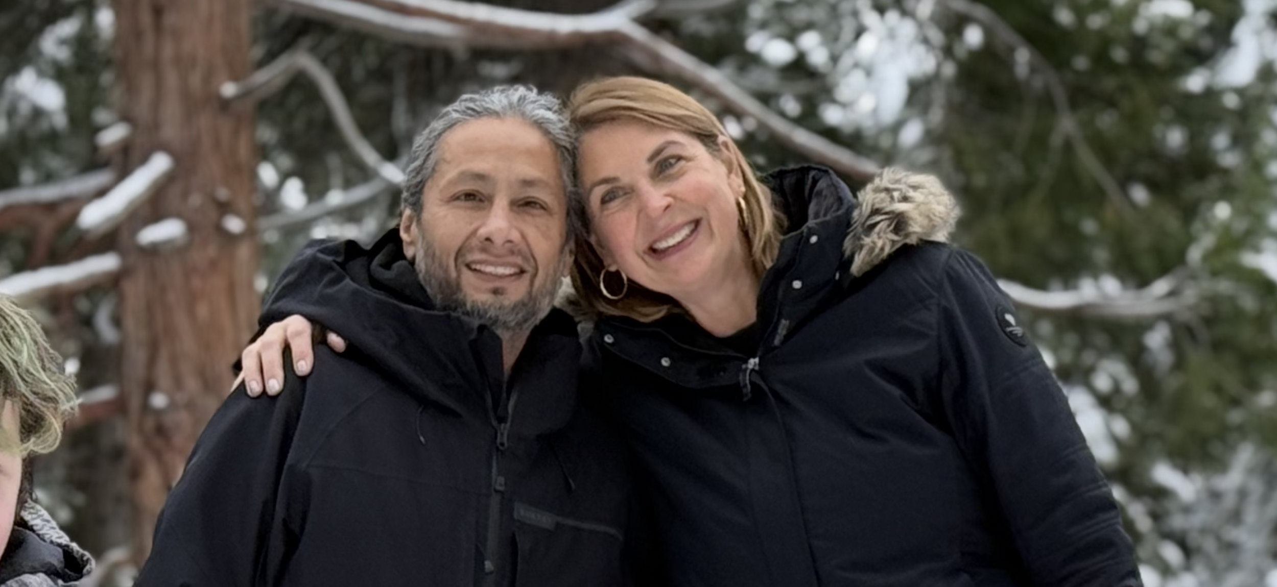Two people smiling and hugging outdoors in a snowy forest, wearing dark winter jackets.
