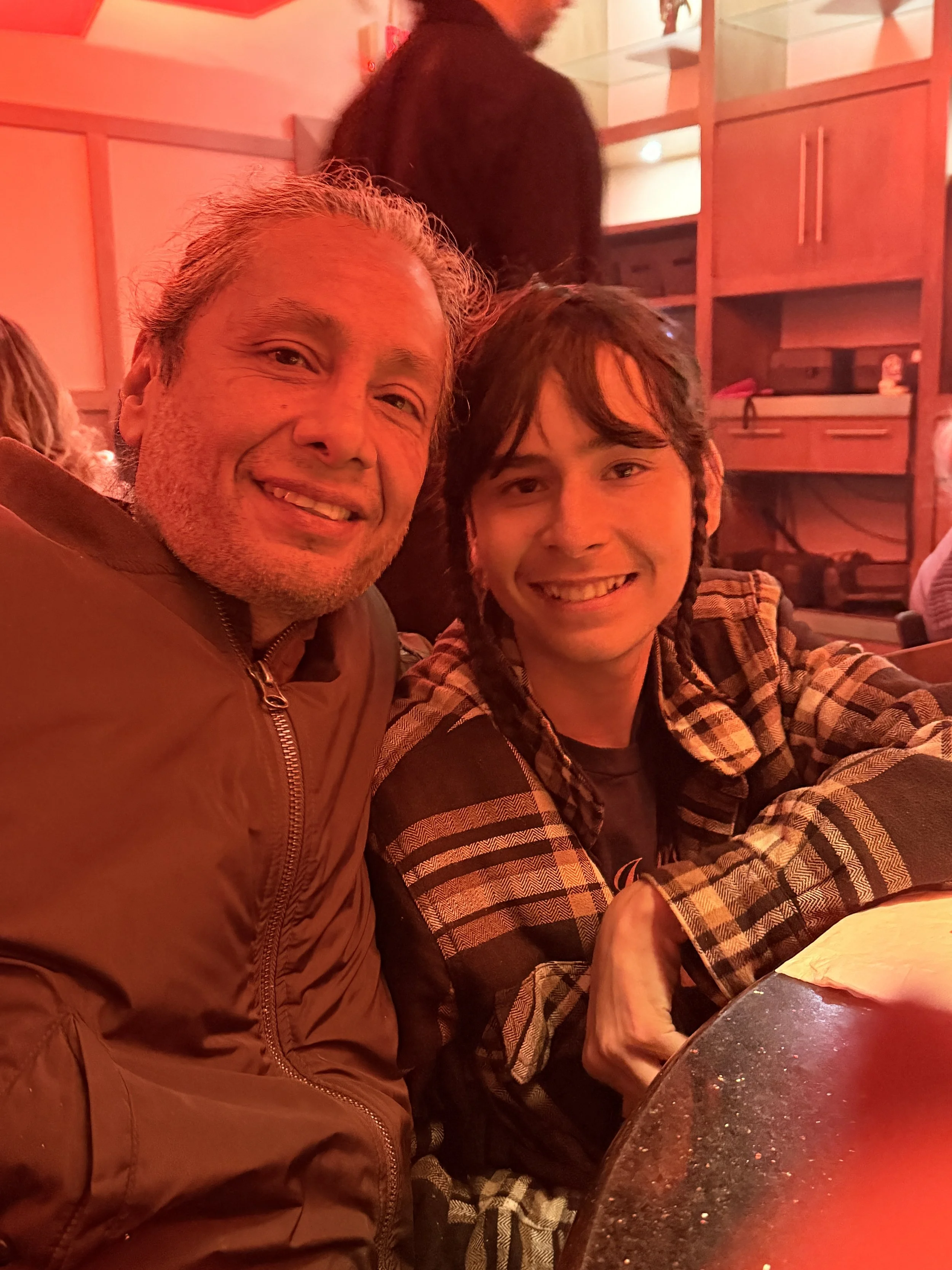 A man and a young man with long hair and braids smiling at a table in a restaurant with red lighting.