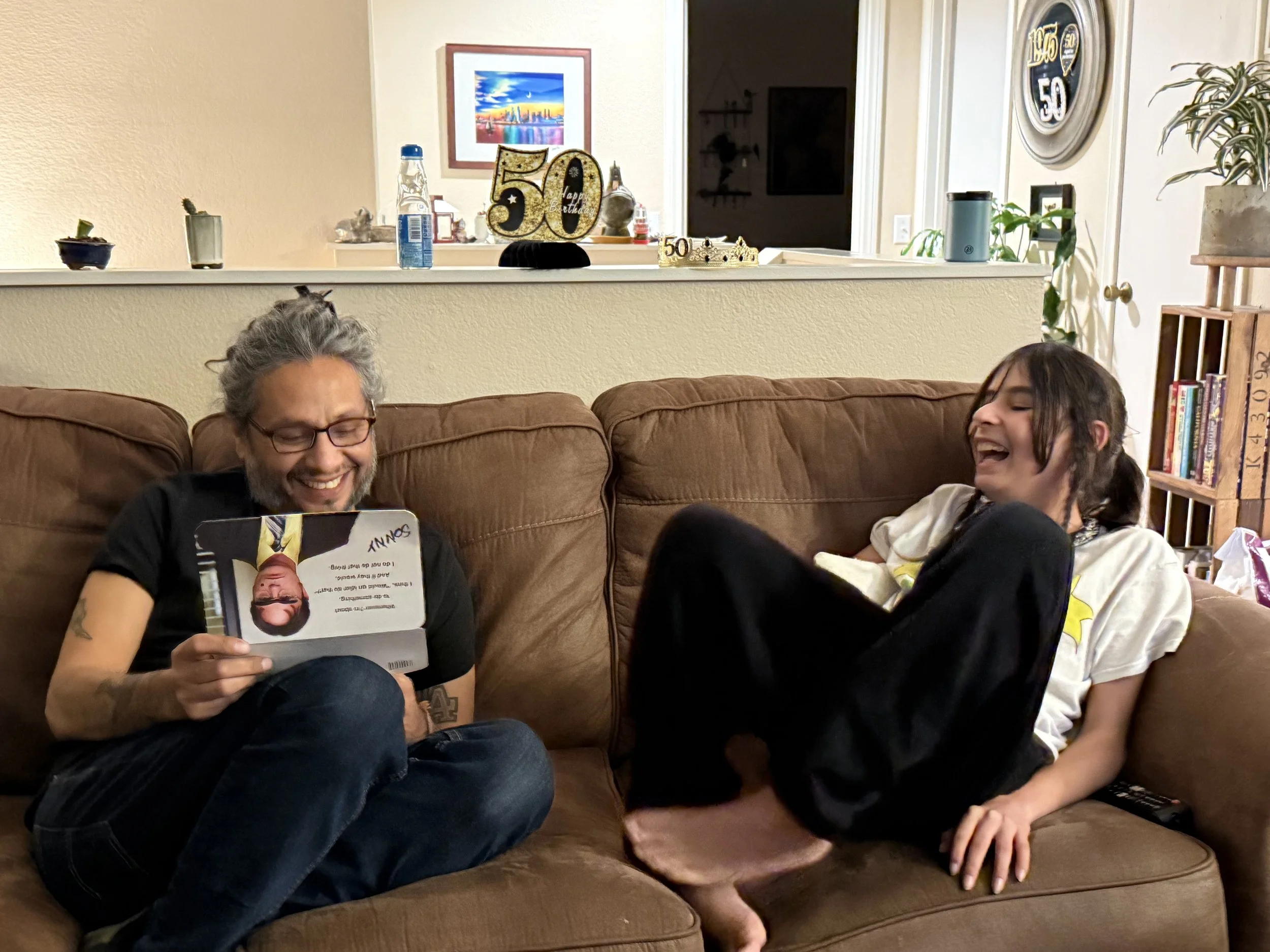 A man with glasses and gray hair sitting on a brown couch, smiling while looking at a card, with a young girl with dark hair sitting on the same couch, laughing and leaning back with her feet up, in a living room decorated with birthday decorations a
