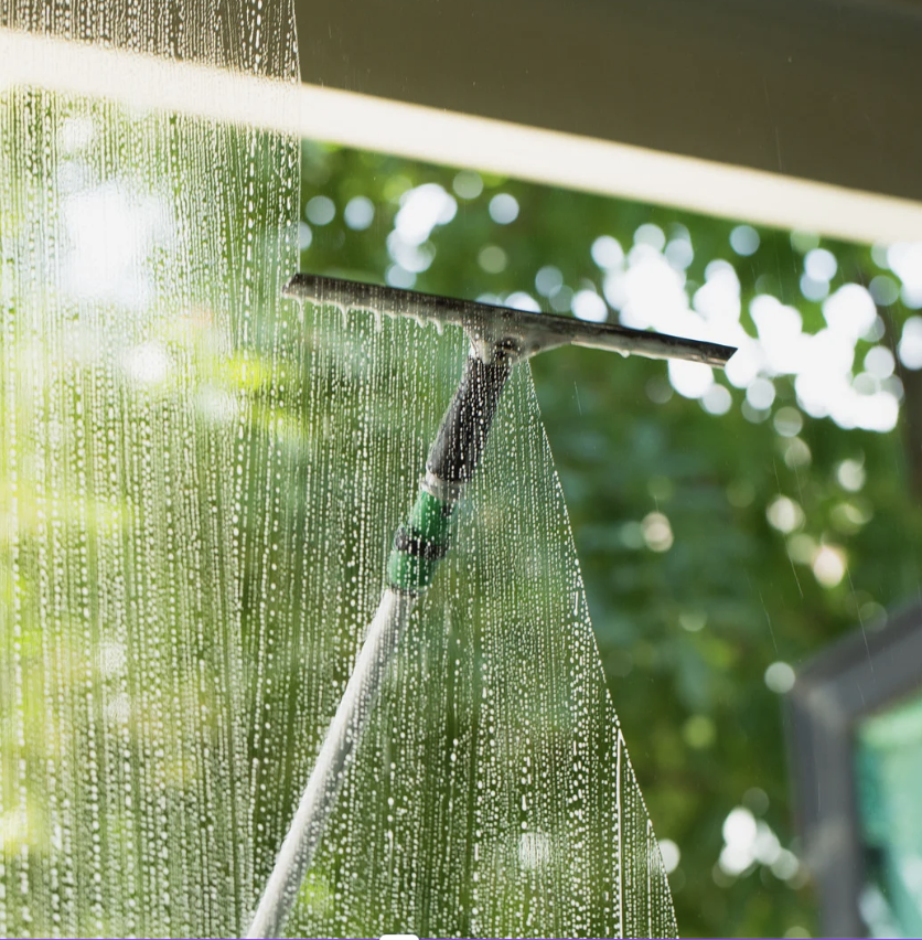 A squeegee with a green handle cleaning a glass window with water droplets, with blurred green foliage in the background.