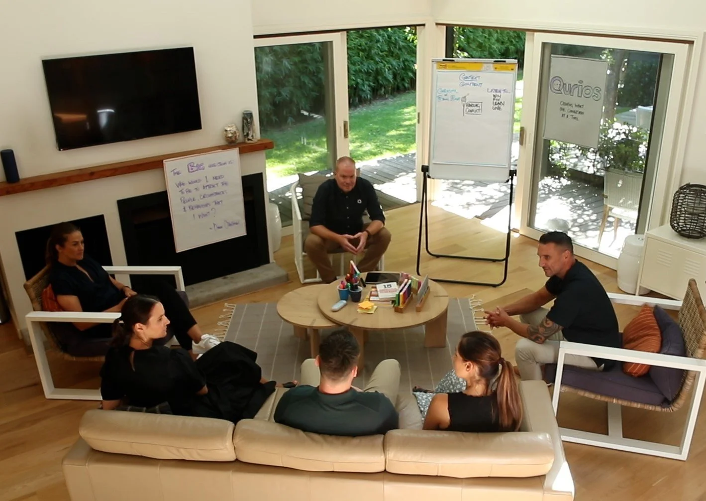 A group of six people sitting in a semi-circle around a round wooden coffee table, in a bright room with large sliding glass doors leading outside, engaged in a meeting or discussion.