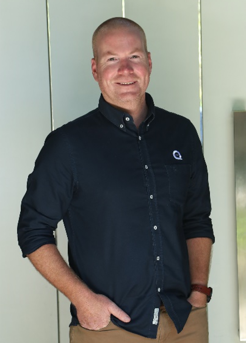 A smiling man with a shaved head, wearing a dark button-up shirt and khaki pants, standing indoors against a light background.