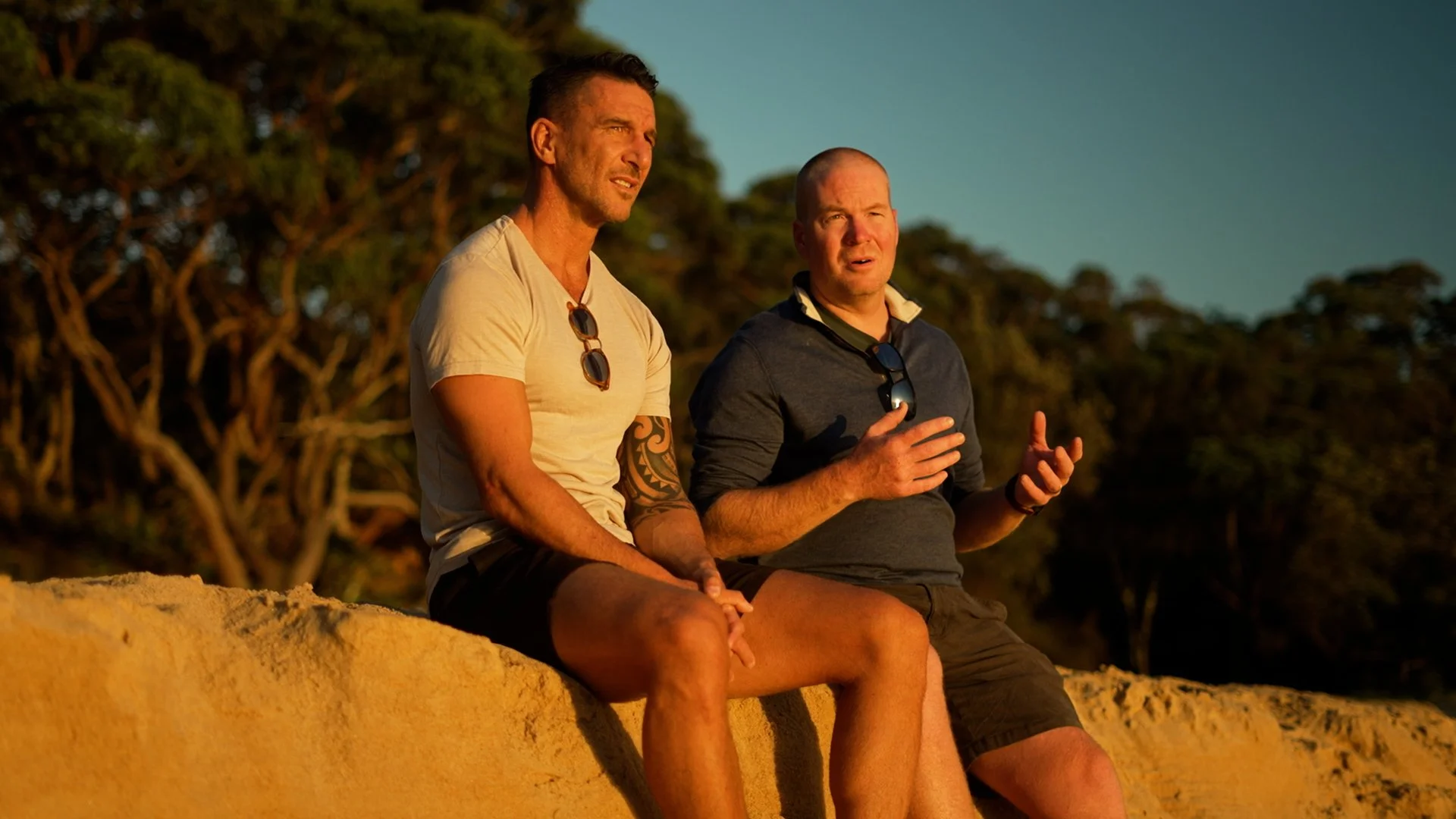Two men sitting on a sandy surface outdoors, engaging in conversation during sunset with trees in the background.