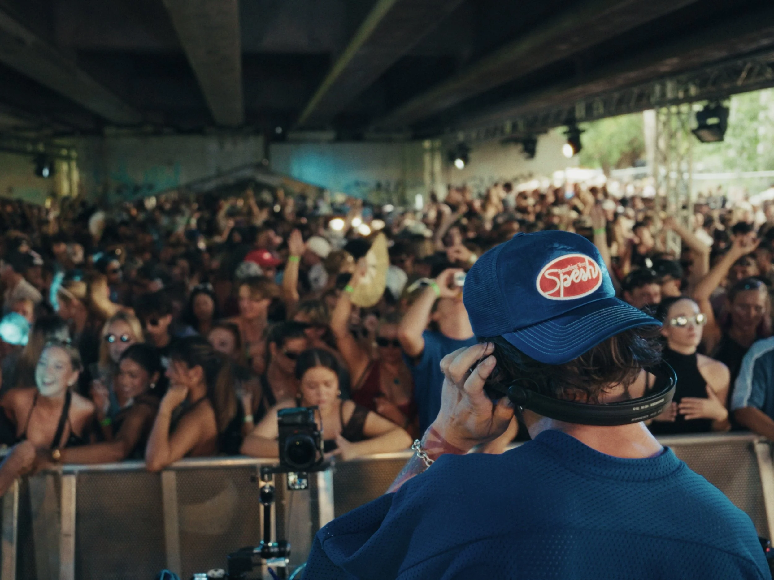 A DJ wearing a blue hat with a 'Speakeasy' logo and headphones, playing music at an indoor music festival with a large, excited crowd in front.