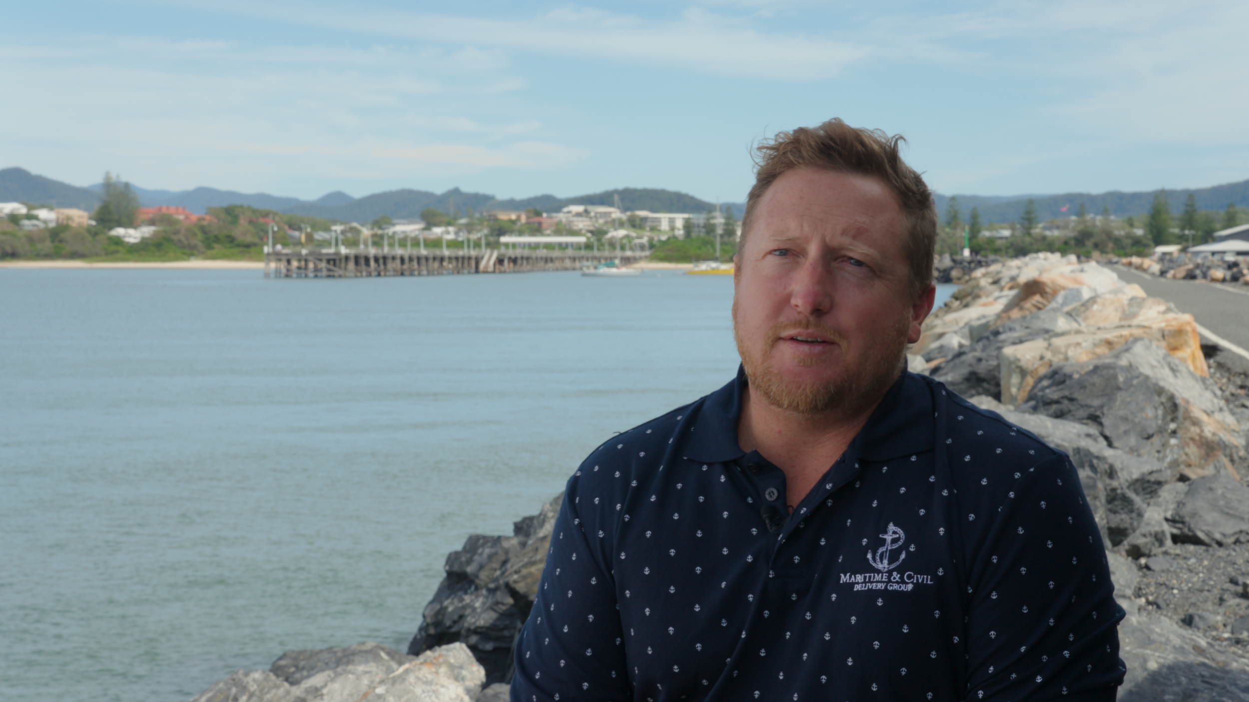 A man with a beard and short hair sitting on rocks near a body of water with a dock and buildings in the background, under a partly cloudy sky.