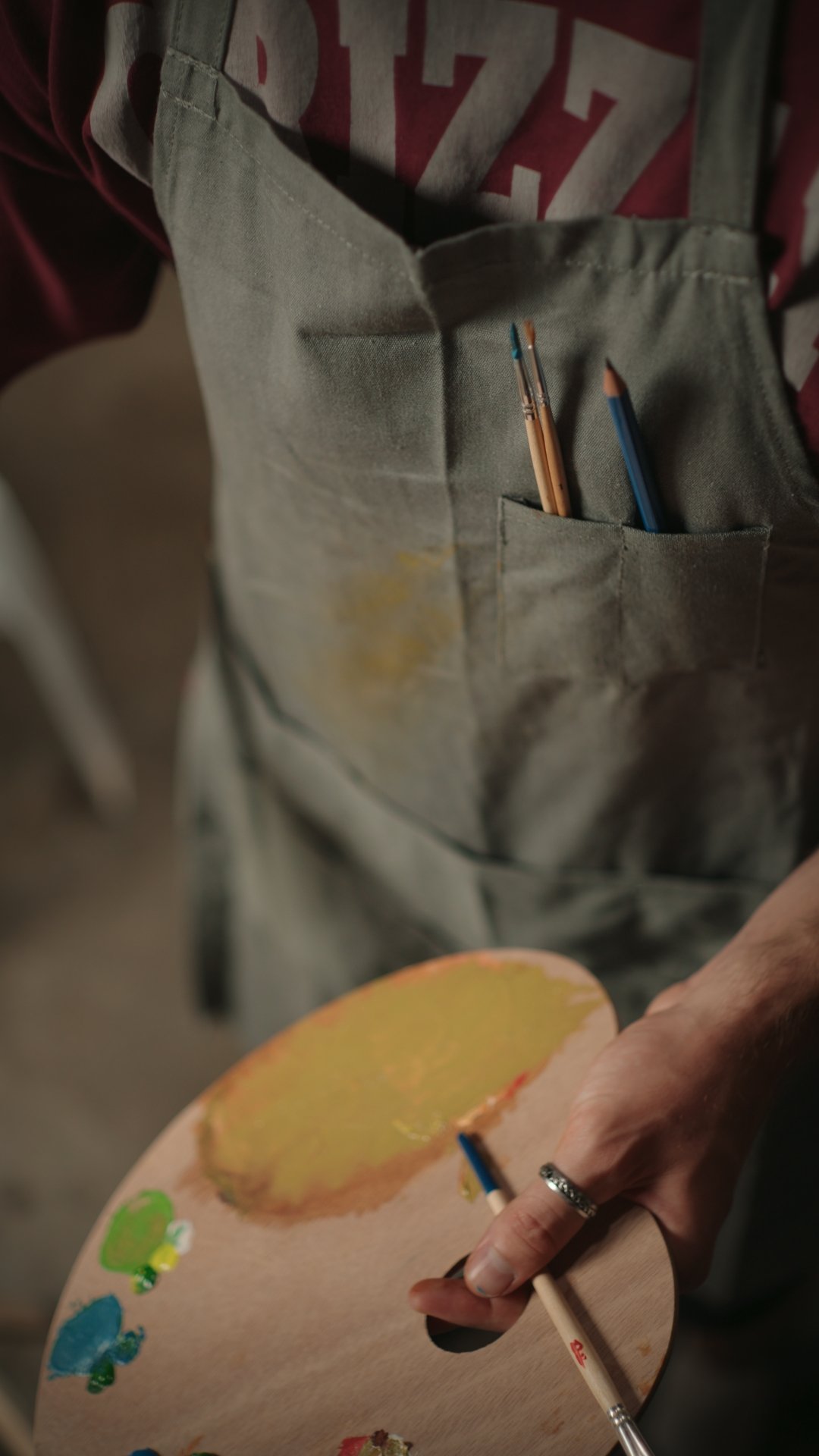 Close-up of a person holding a paint palette with colorful paint blobs, wearing a ring, with a gray apron with paint stains and two brushes tucked into a pocket.