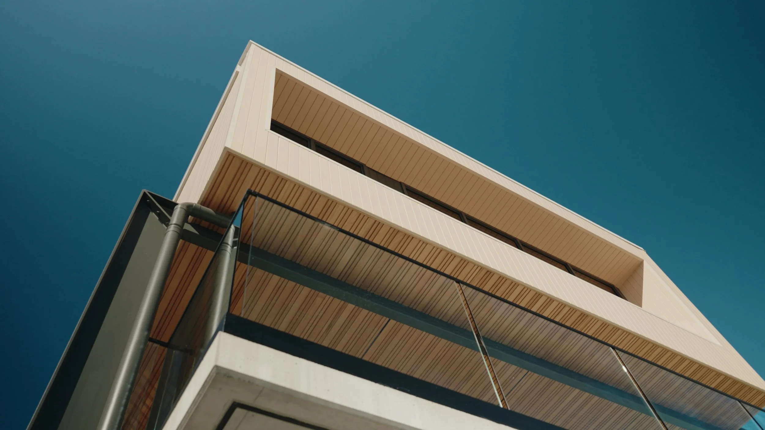 Low-angle view of a modern building with beige siding, large glass balcony with black railings, and a clear blue sky in the background.