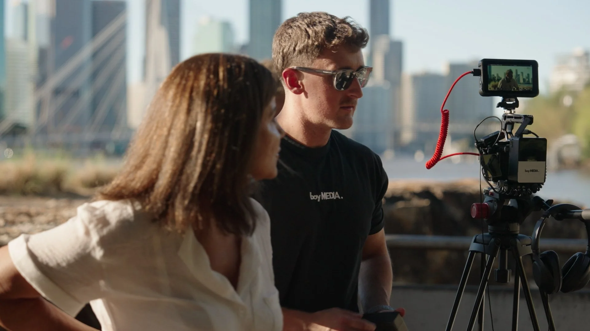 Two people, a woman and a man, are filming outdoors near a city skyline. The man is wearing sunglasses and a black t-shirt, while the woman has shoulder-length hair and is looking away. A professional camera on a tripod is filming them.