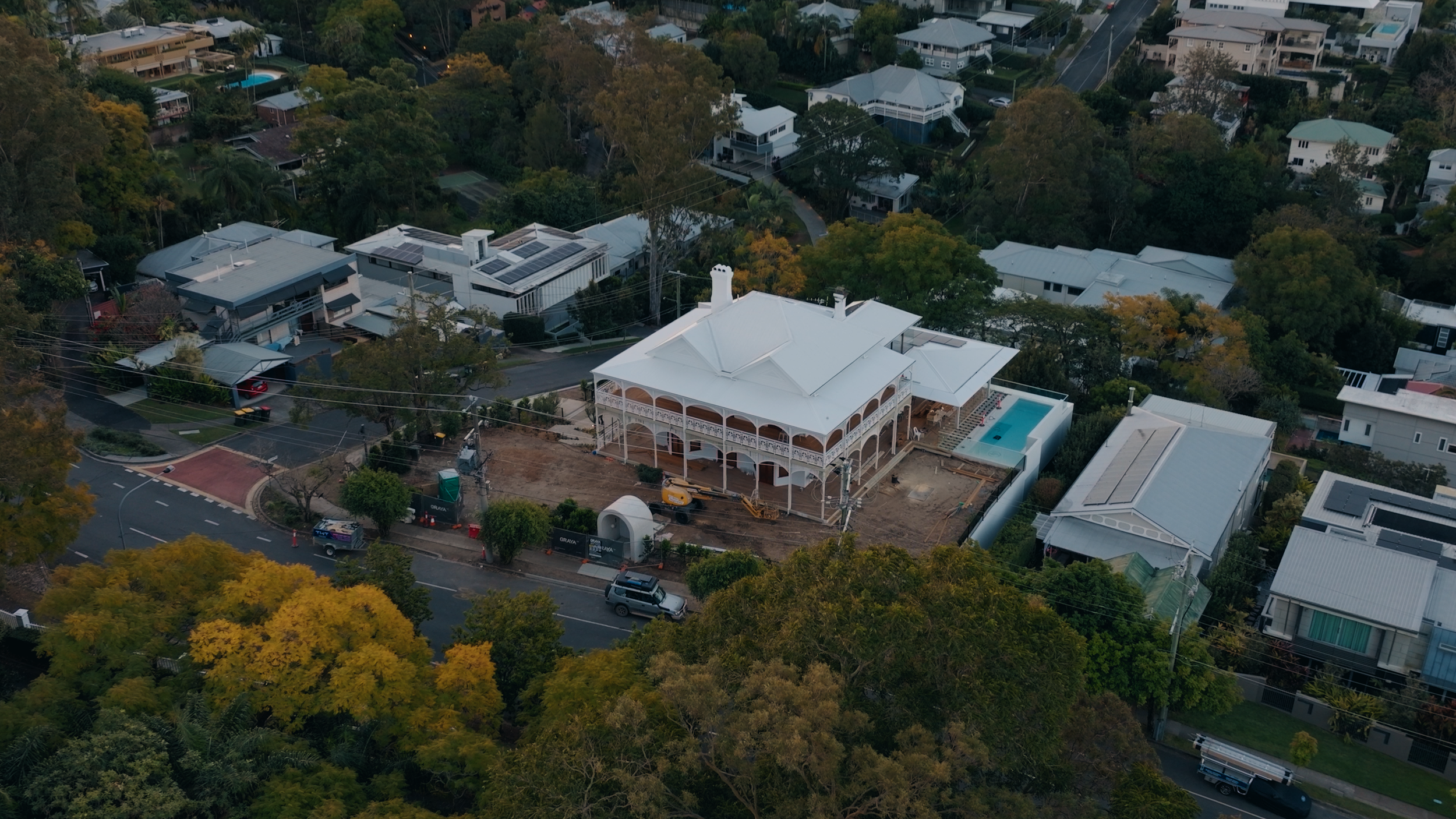 Aerial view of a large, Victorian-style house under construction in a residential neighborhood, with a white exterior and pool nearby, surrounded by trees and other houses.