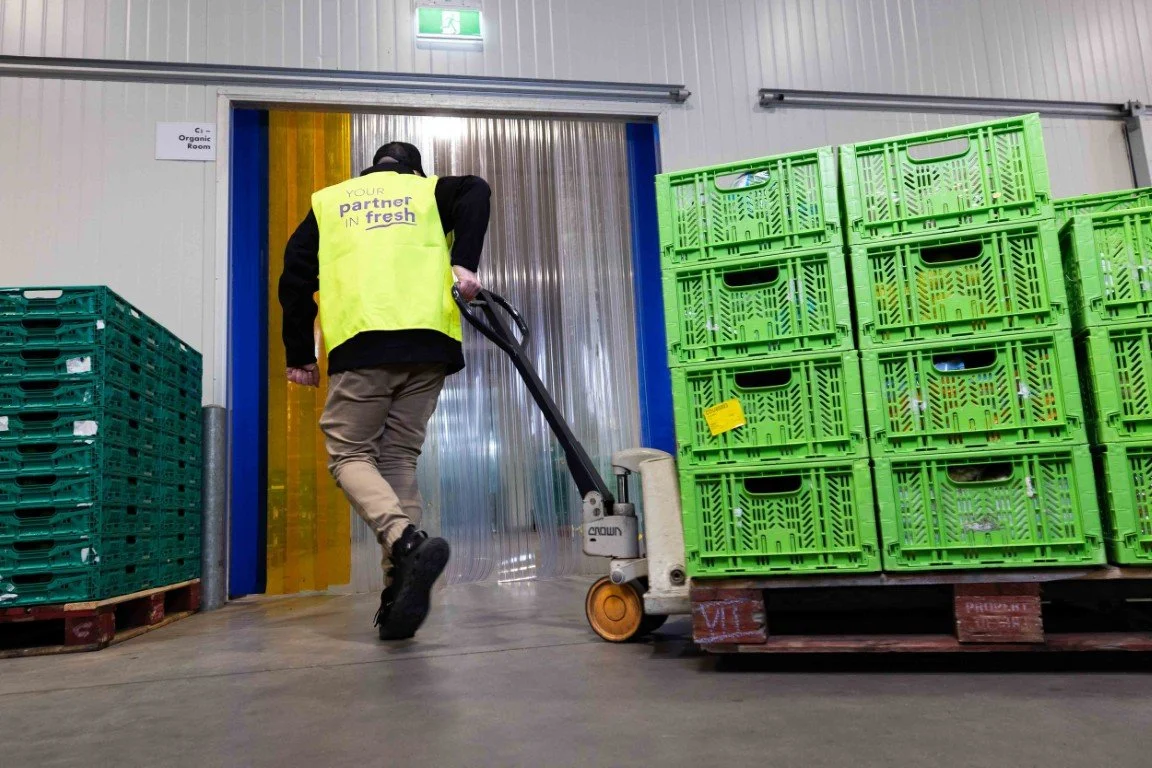 A worker in a yellow safety vest pushes a hand pallet truck loaded with green plastic crates inside a warehouse or storage facility.