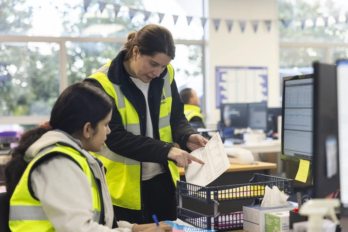 Two women working together in an office, both wearing yellow safety vests, with one woman showing a document to the other.