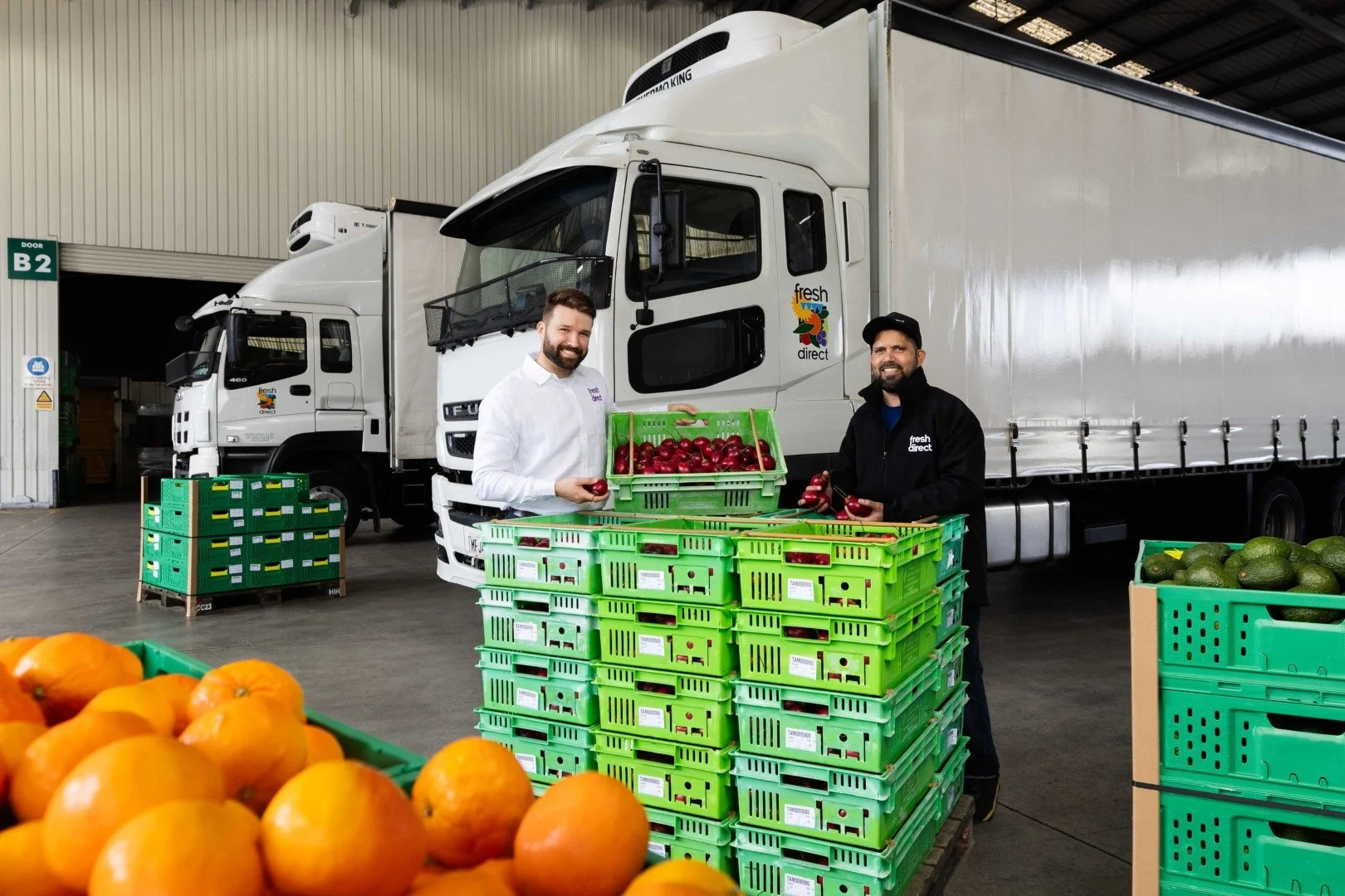 Two men standing inside a warehouse next to green crates filled with red apples and avocados. The men are smiling and holding apples, with trucks and more produce in the background.