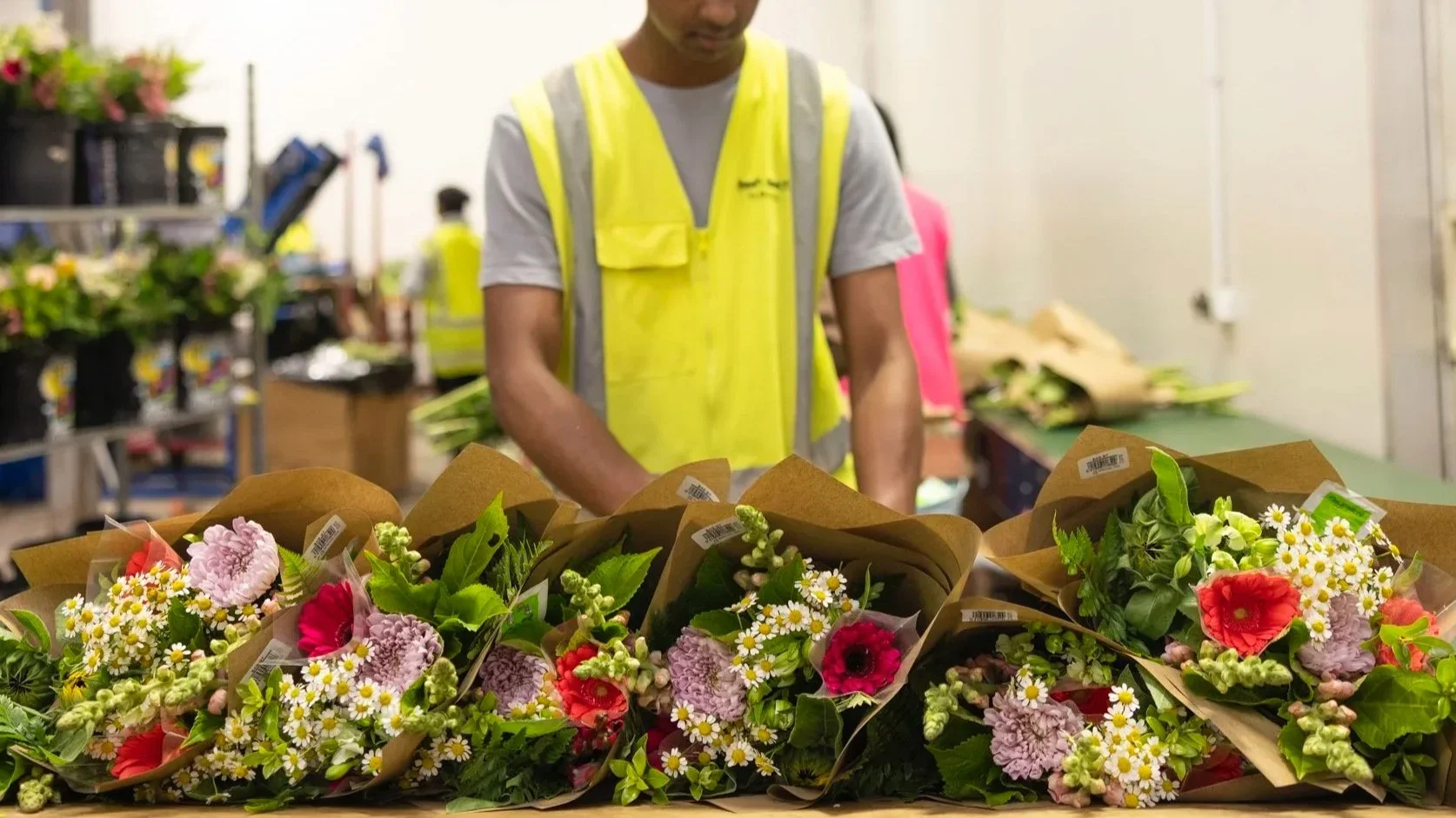 A person in a yellow safety vest arranging bouquets of colorful flowers on a table in a flower shop or warehouse.