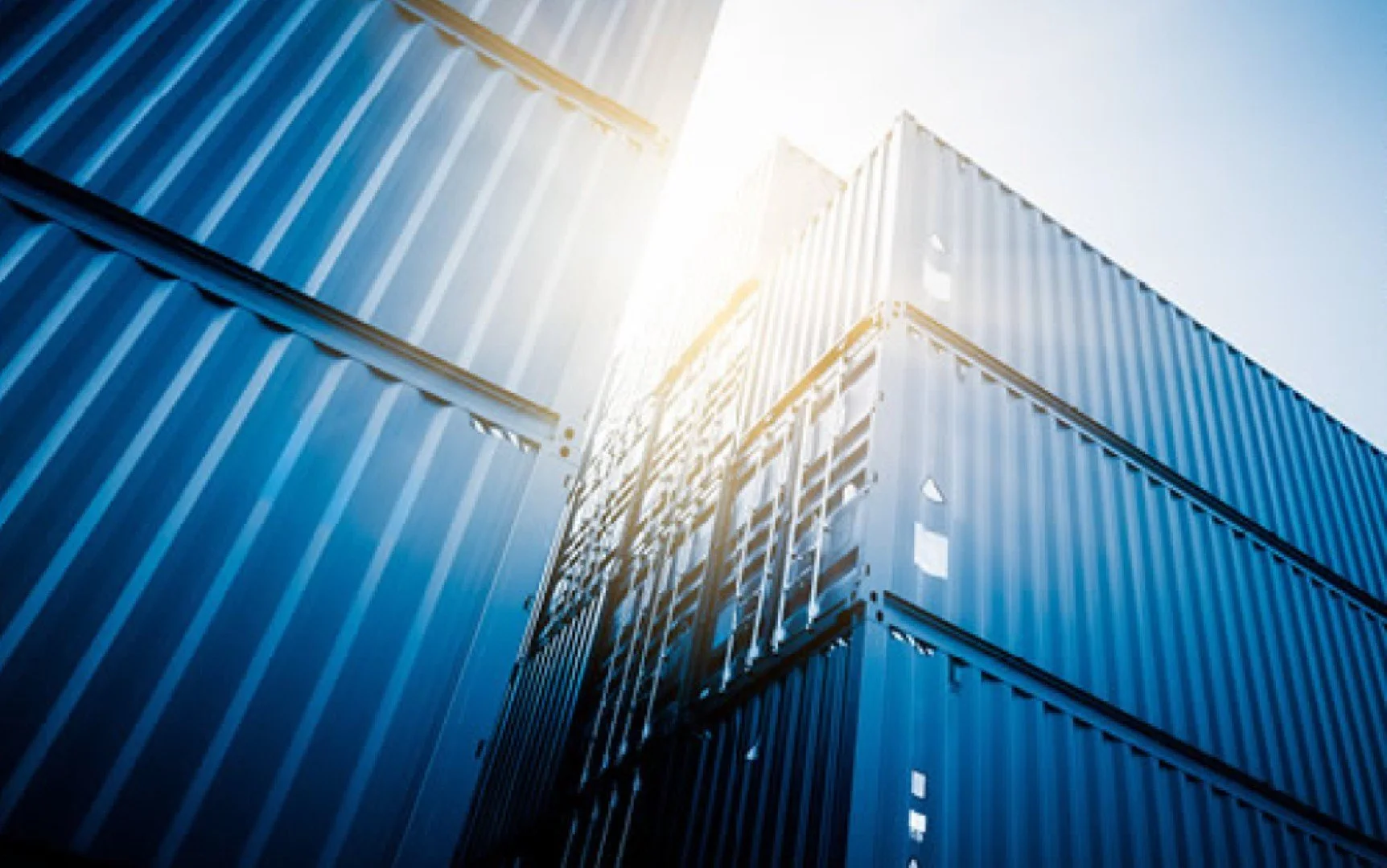 Upward view of stacked blue shipping containers with sunlight in the background.