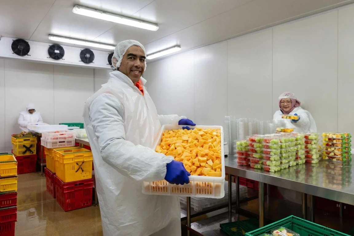 Workers in protective clothing packing slices of melon and other fruits in a clean food processing facility.