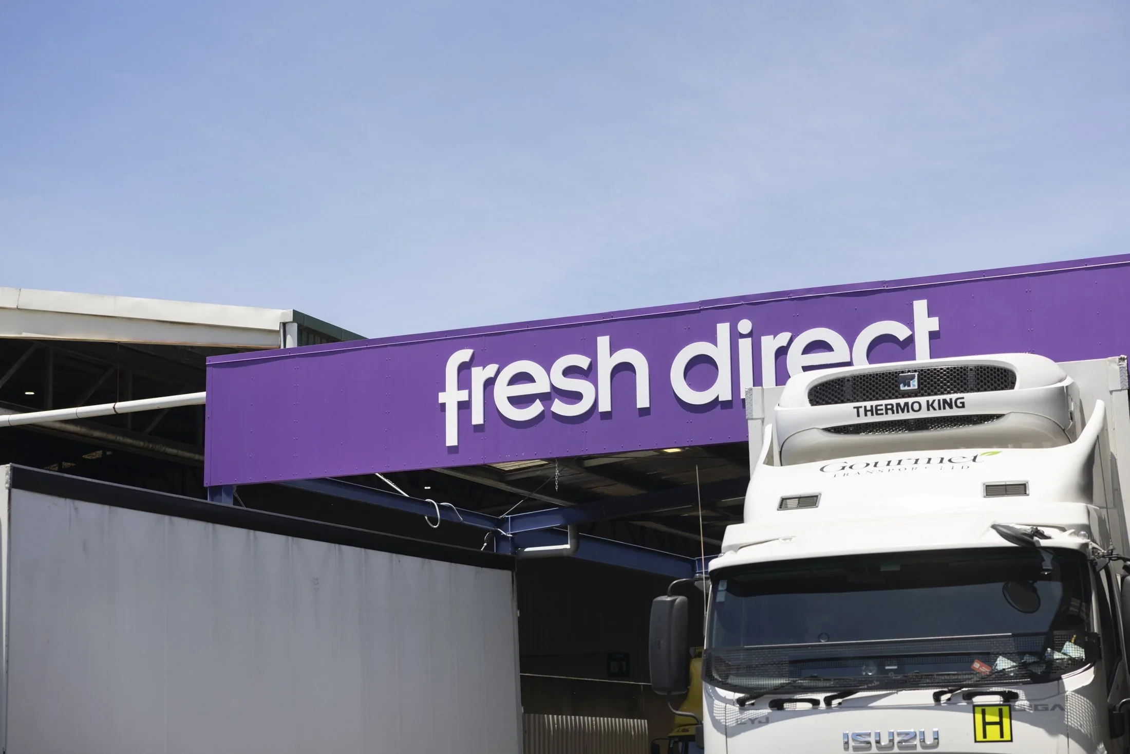 A purple signboard with white text that reads 'fresh direct' is mounted on a building. Part of a white truck with an Isuzu logo and a Thermo King refrigeration unit is visible in front of the building. The sky is clear and blue.