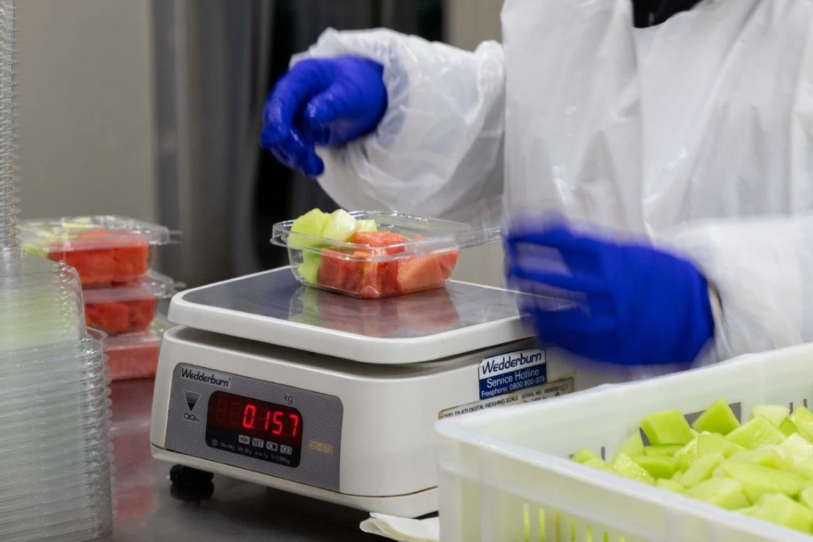 Person wearing blue gloves and a white apron weighing cut pieces of watermelon and honeydew on a digital kitchen scale.
