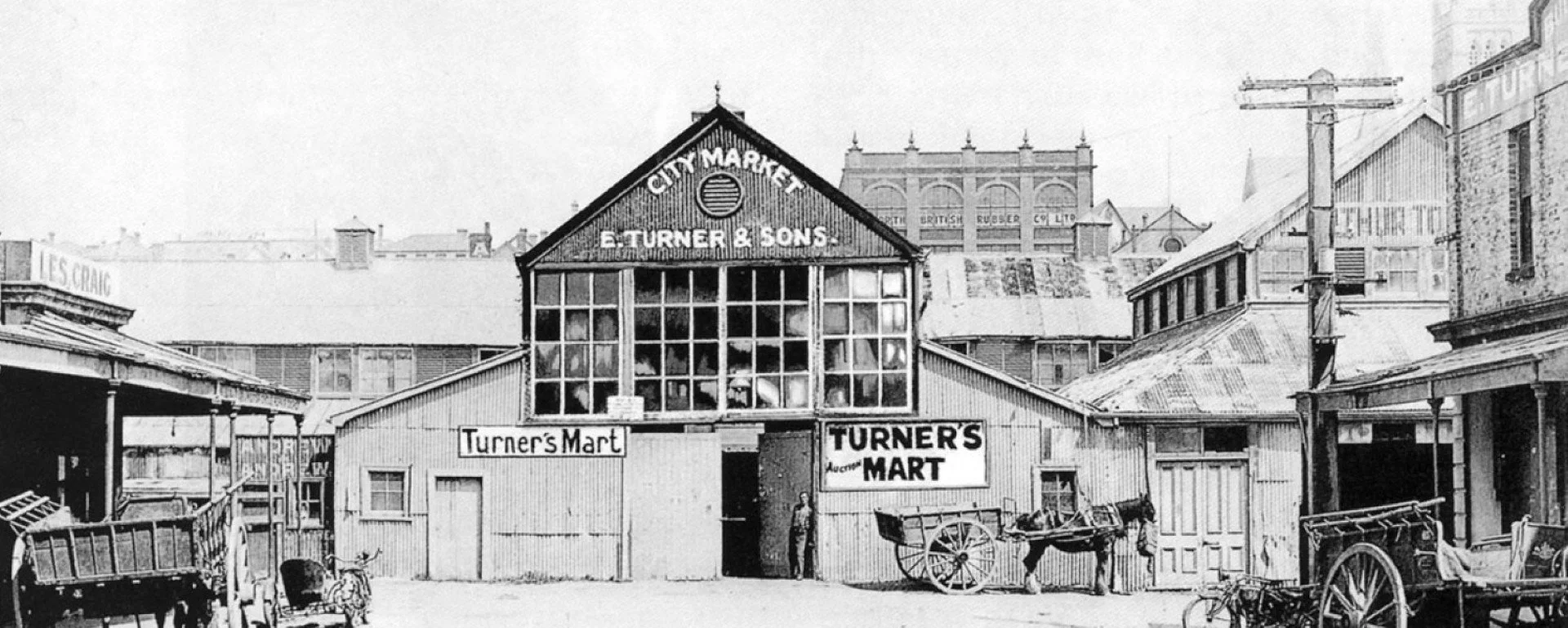 Historical black and white photograph of Turner’s Mart, a market building with a sign reading "City Market" and "Turner & Sons." The building has large windows and outdoor carts with people standing nearby. Surrounding buildings are also visible.