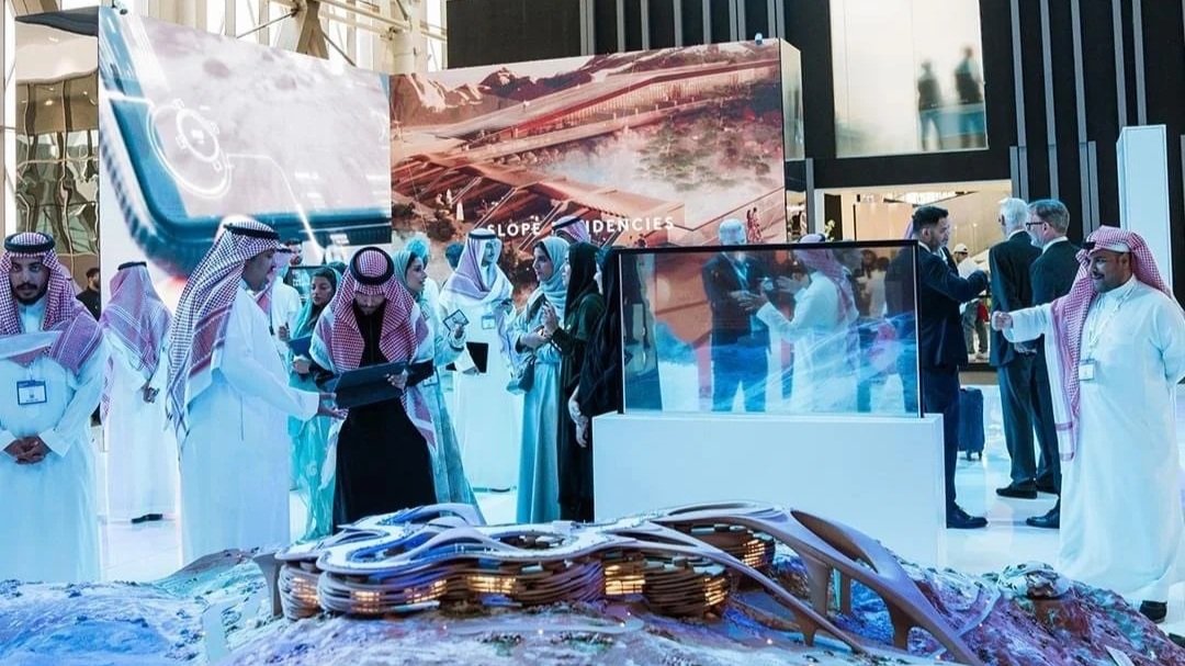 Group of Middle Eastern people in traditional attire and business suits gathered around technology exhibits at a conference or expo, with digital displays and large posters in the background.