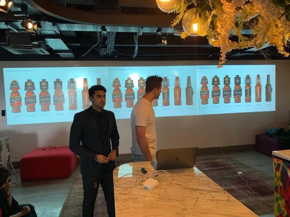 Two men standing in front of a large screen displaying images of beer bottles in a conference or presentation room, with some indoor lighting and decorations hanging from the ceiling.